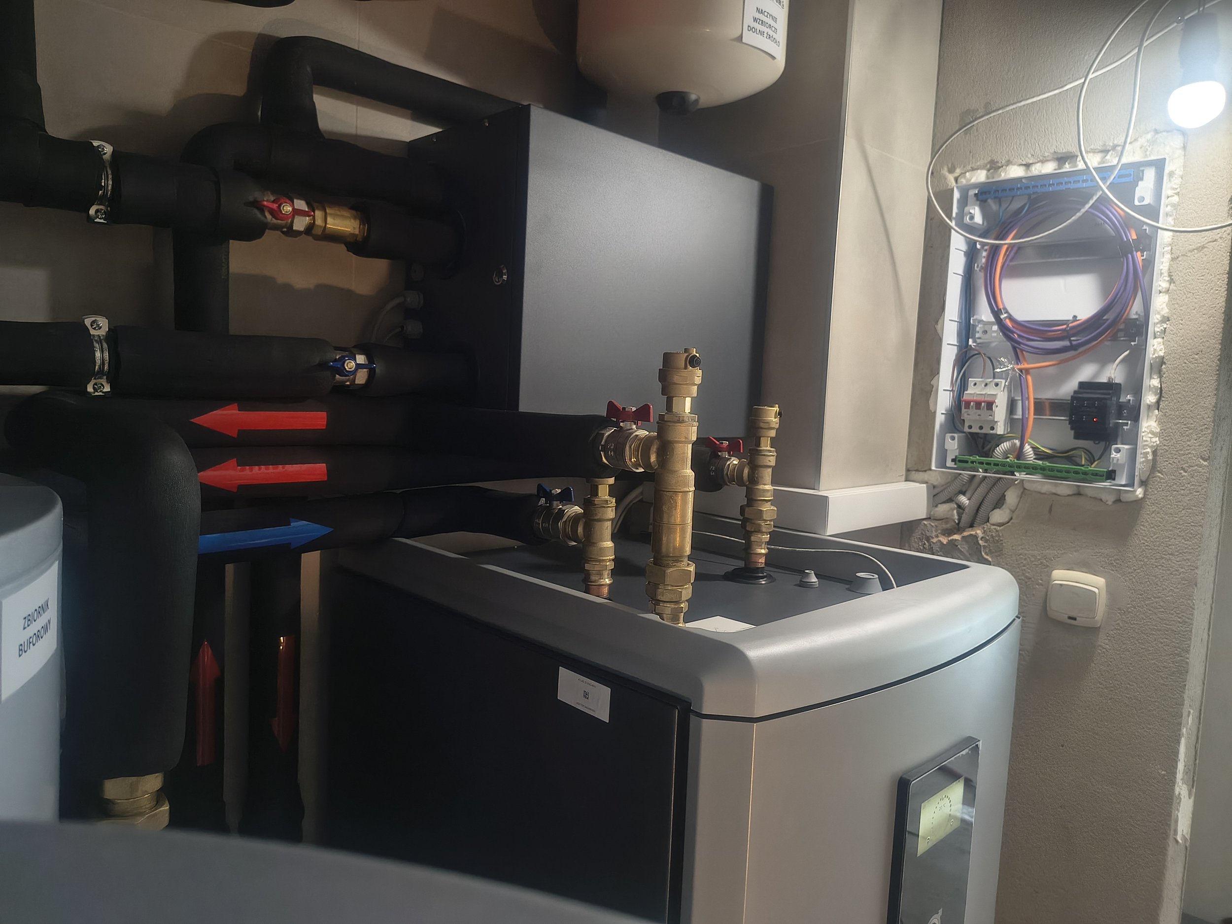 A space with plumbing and electrical components, showing black pipes with red and blue valves, a brass valve, a white panel with exposed wiring, and a water heater tank.