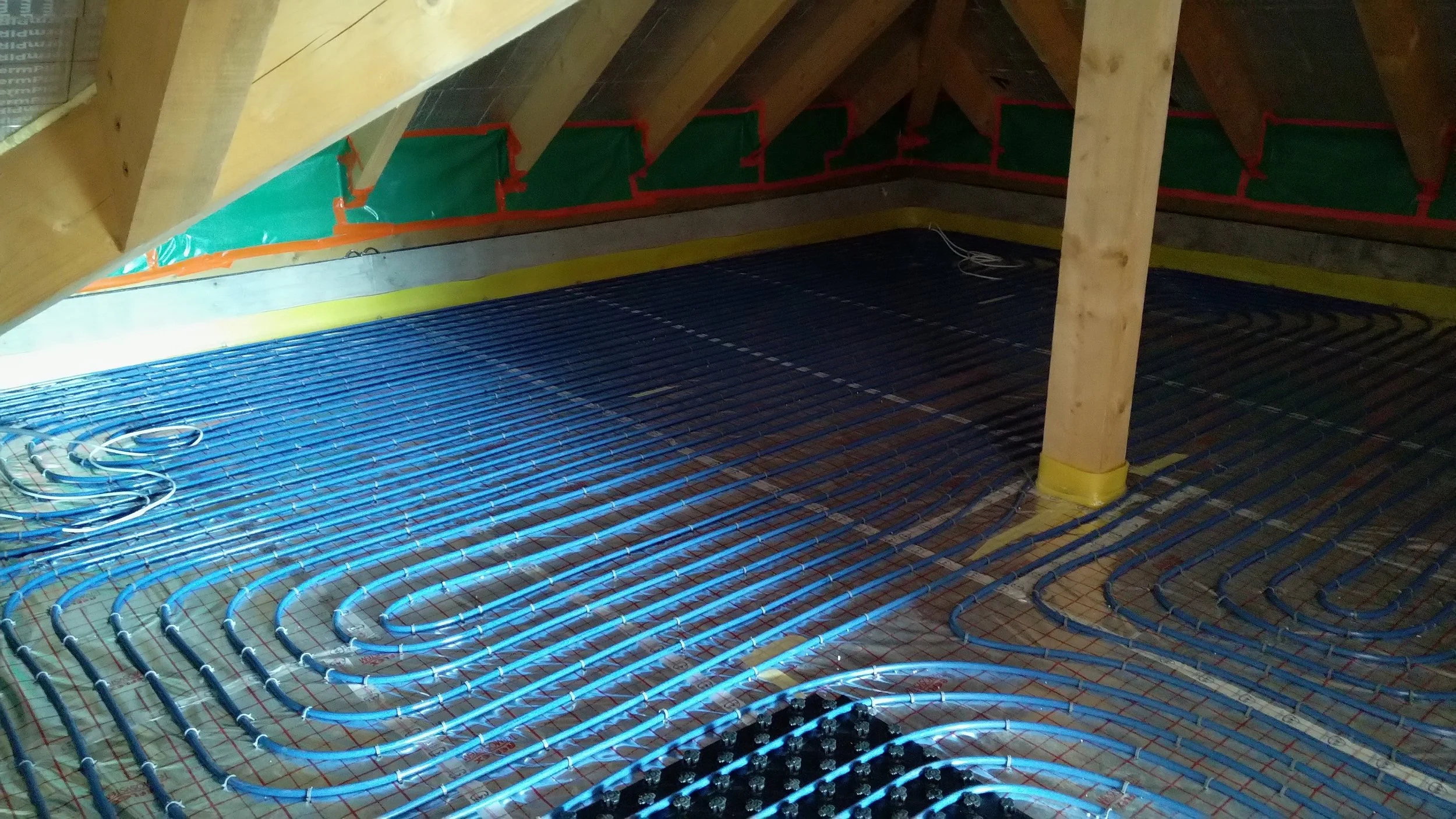 Installation of radiant floor heating with blue heating cables laid over a concrete floor, with wooden roof rafters visible above.