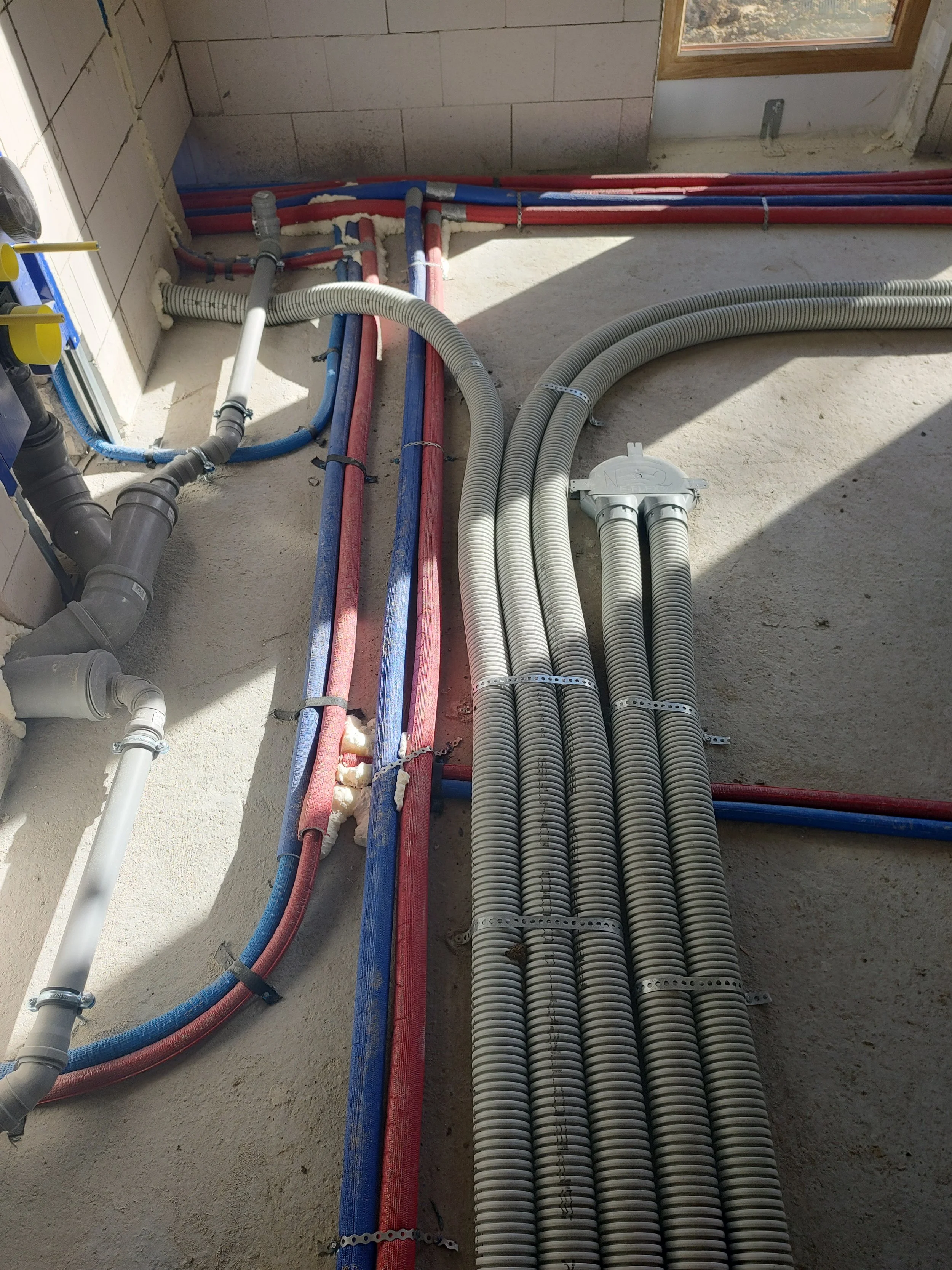 Interior view of a construction site showing organized electrical and plumbing pipes and conduits on a concrete floor, with a window on the right and unfinished wall surfaces.