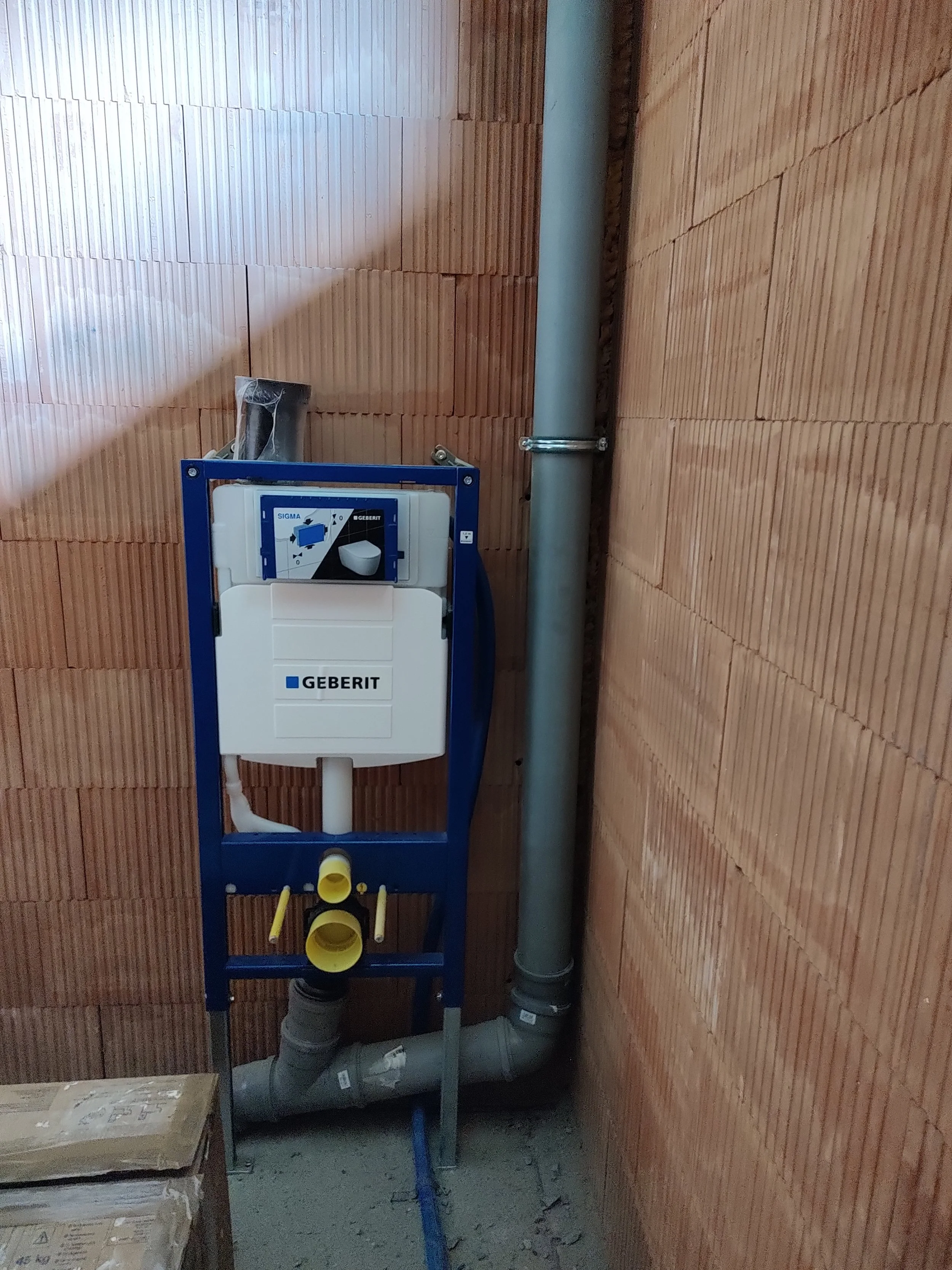 An in-progress toilet installation in a room with unfinished brick walls. The toilet's tank is in a blue metal frame, and several pipes and plumbing components are visible. There are cardboard boxes on the floor.