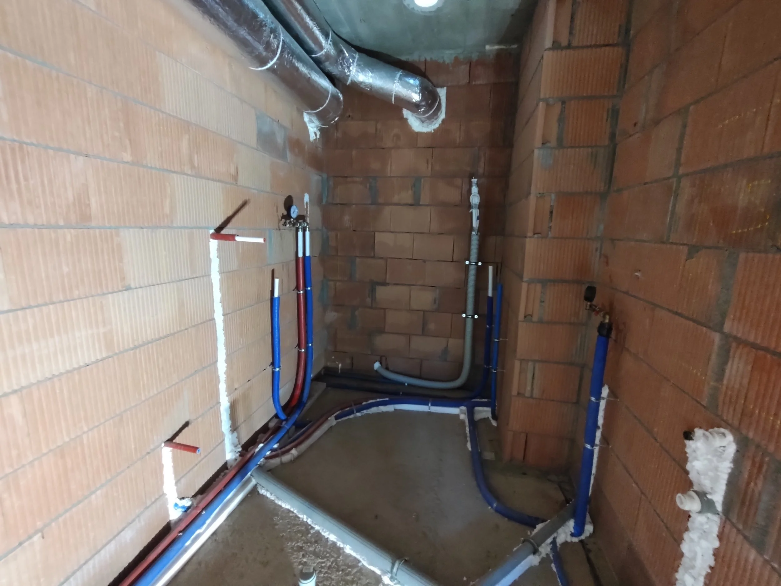Plumbing pipes installed inside a brick-walled room, with various blue and red pipes, and some insulation foam around fittings.