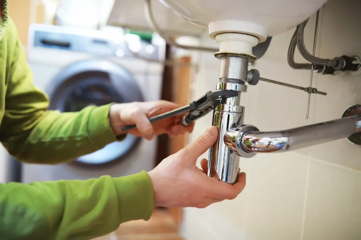 Person working under a kitchen sink, using a wrench to repair or maintain the plumbing.