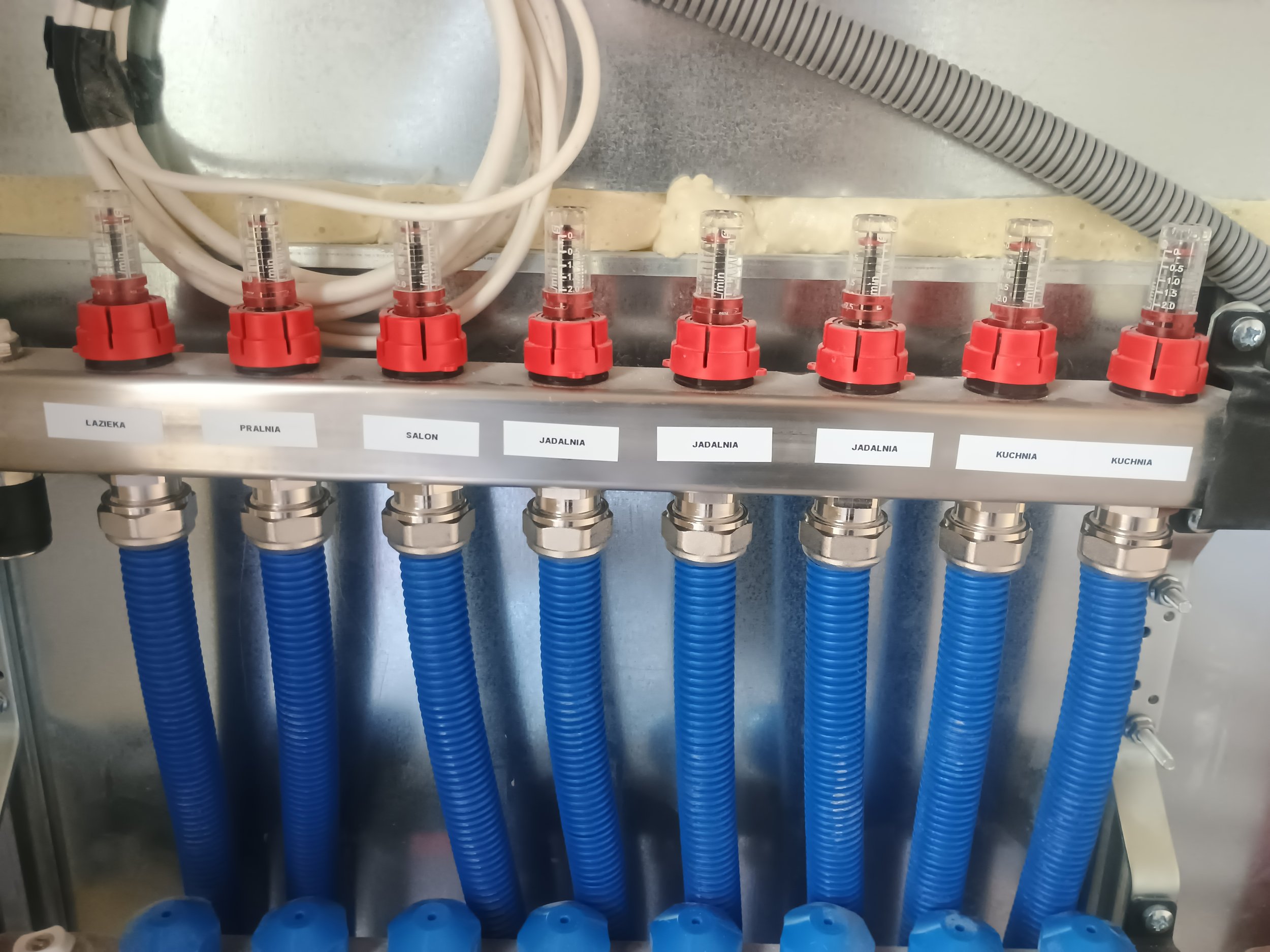 A plumbing manifold with blue flexible hoses connected to it, labeled with areas such as "LAZIENKA," "PRALNIA," "SALON," "JADALNIA," and "KUCHNIA".