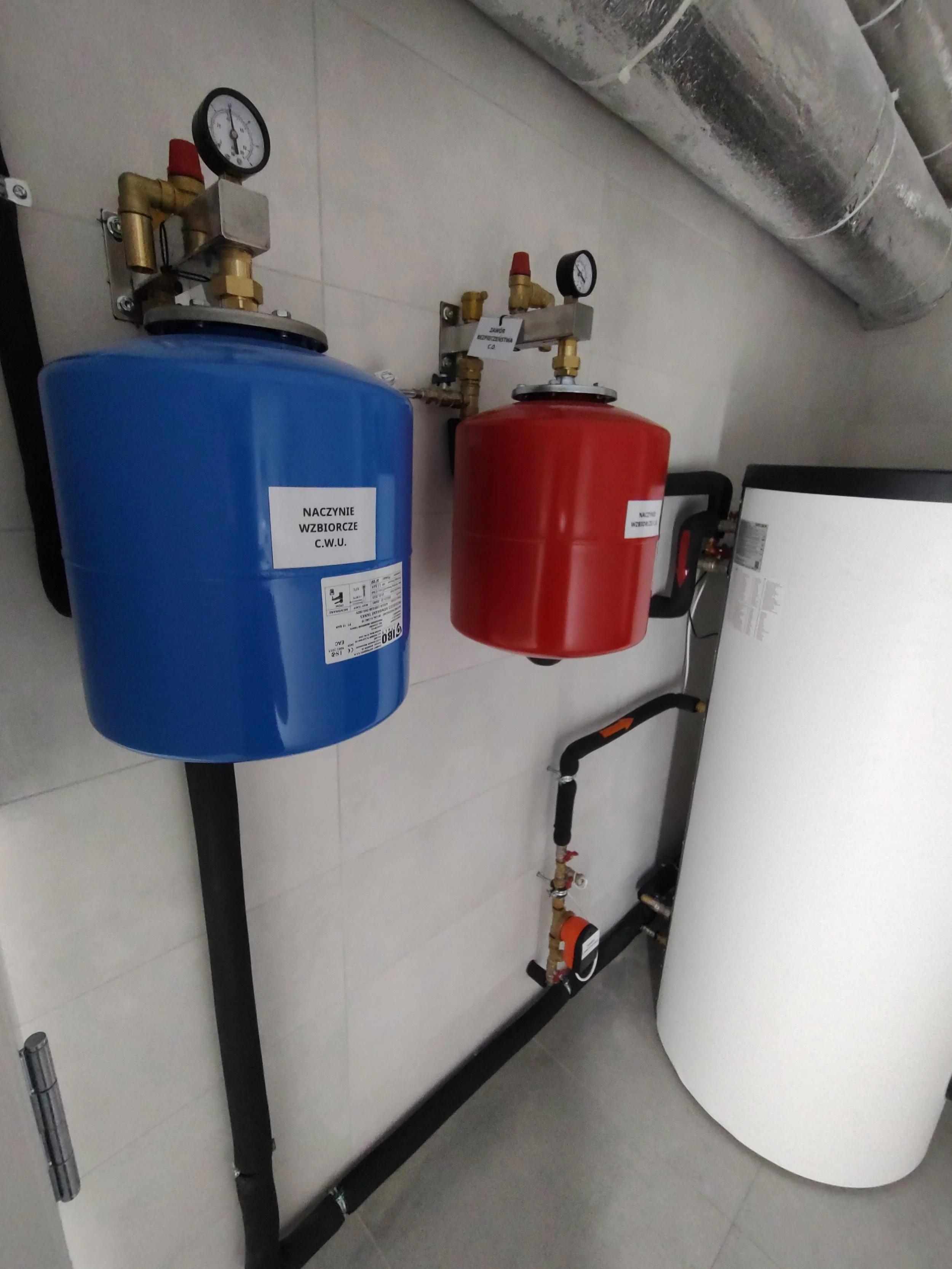 A blue expansion tank and a red expansion tank connected to pipes, gauges, and valves on a wall in a mechanical room.