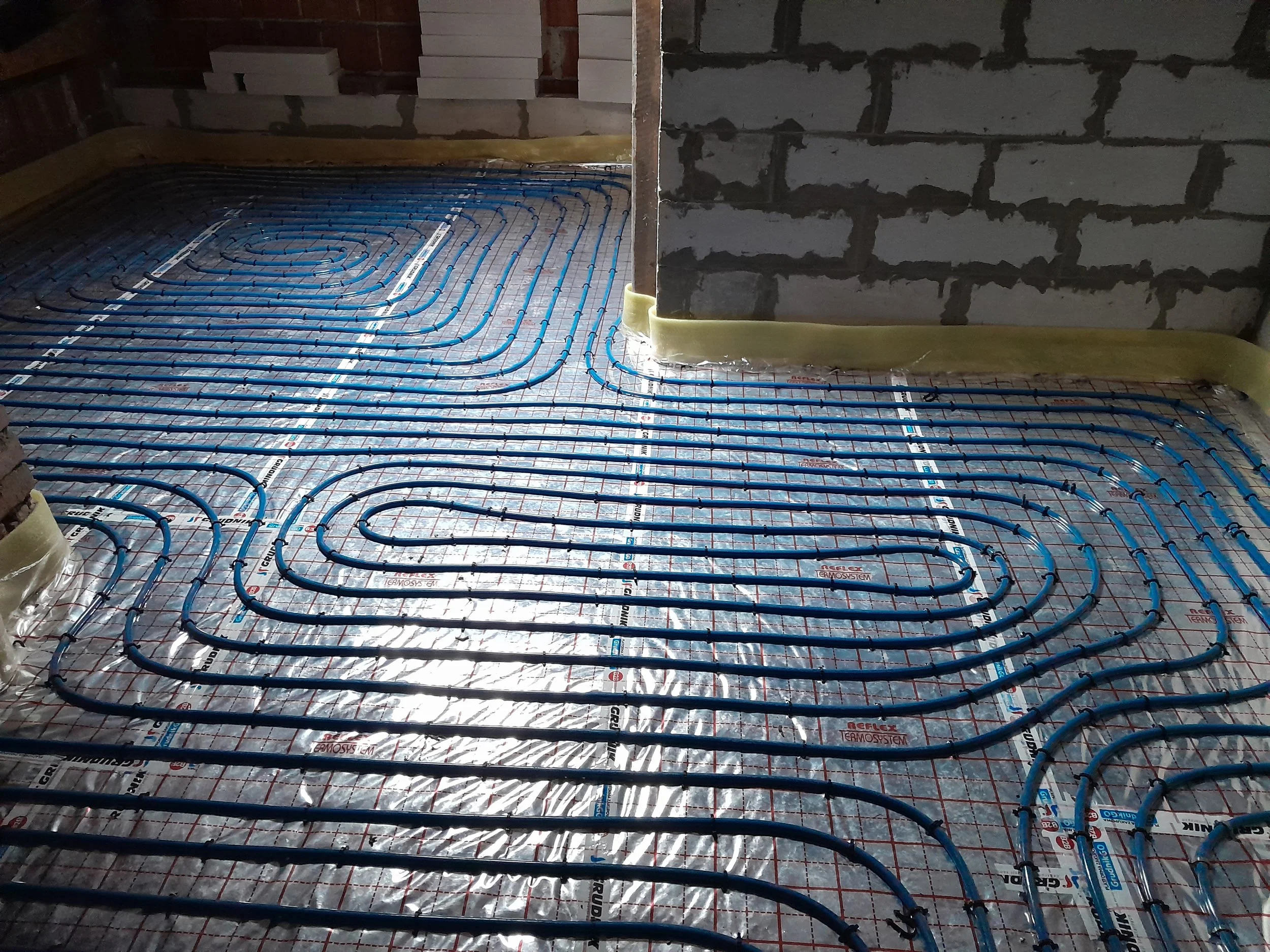 Underfloor heating system with blue pipes installed on a reflective insulation sheet in a room under construction, with a partially built brick wall in the background.