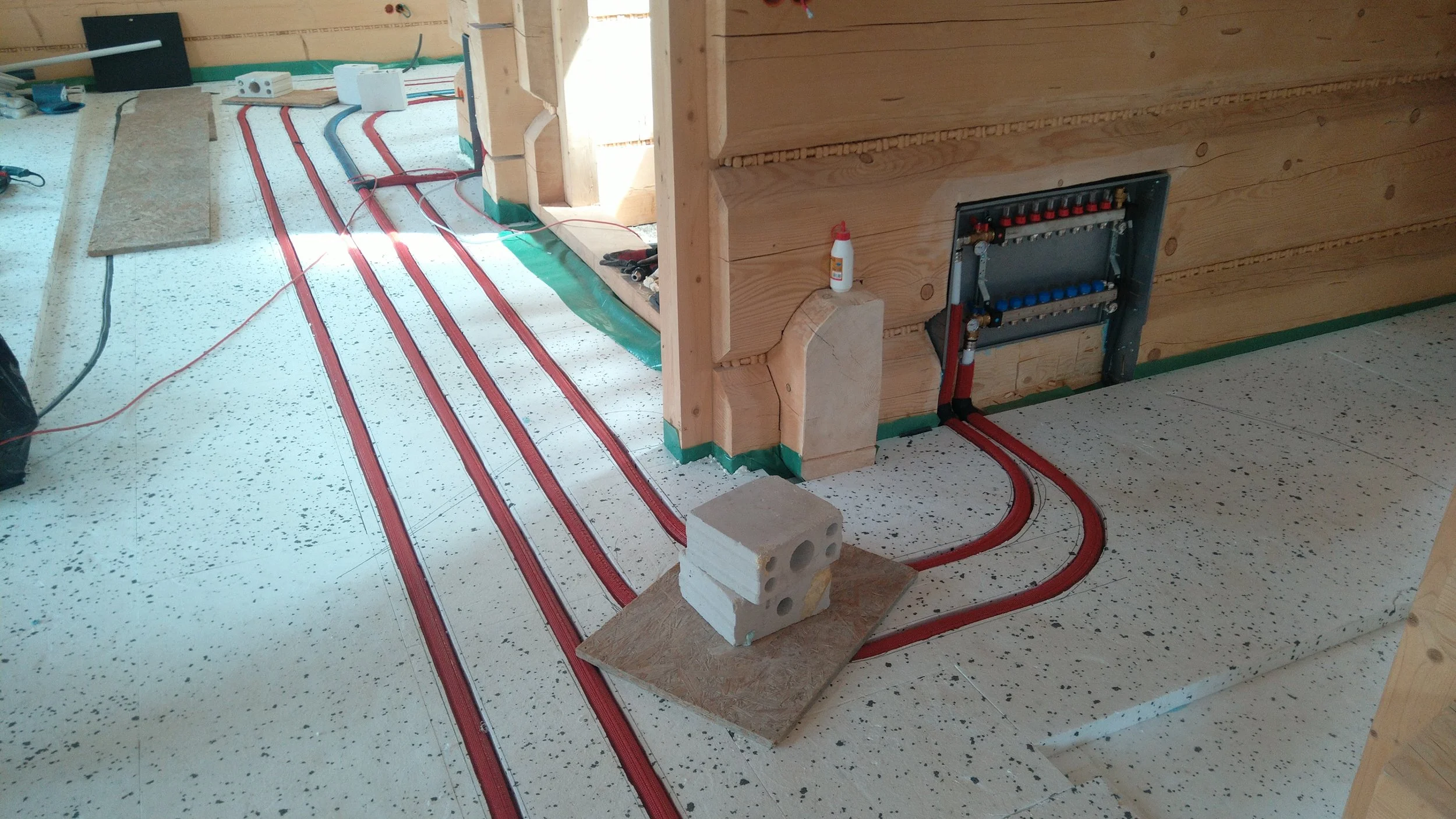 Underground heating or plumbing system installation in a wooden structure, with red and black piping laid on a white speckled floor, construction materials, and tools present.