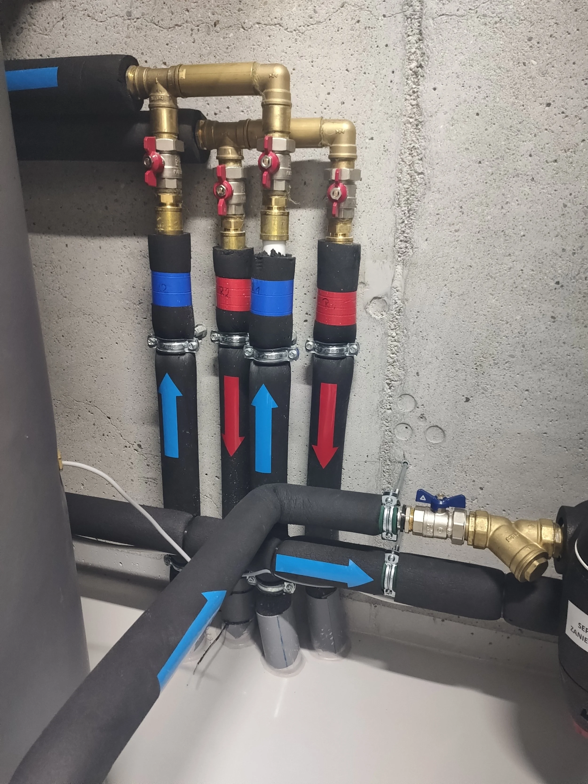 Piping system with black hoses, brass fittings, and color-coded valves on a concrete wall.
