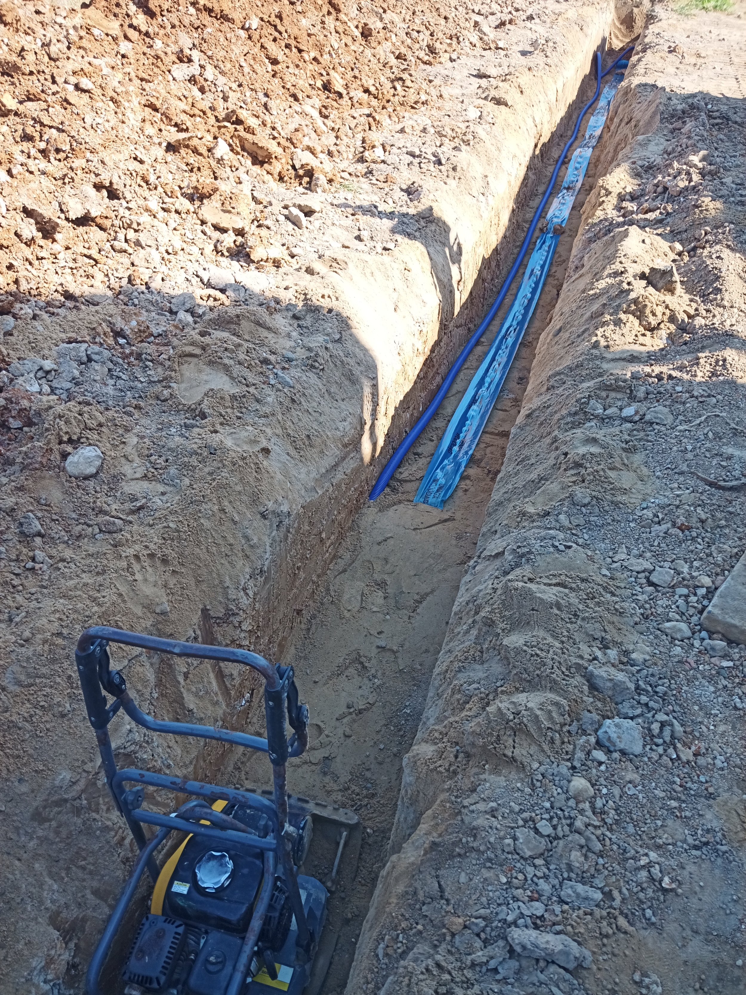 Excavated trench with blue pipes laid inside, construction tools and equipment in the trenches, dirt and rocks surrounding the area.