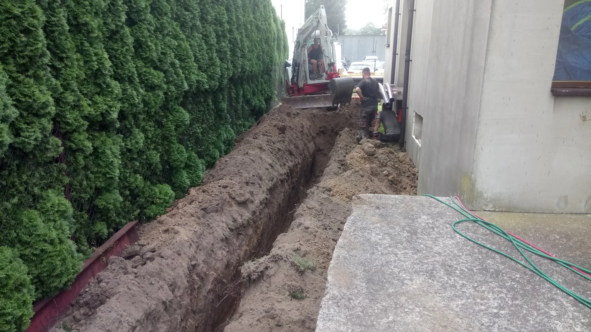 Construction workers doing trench work beside a building with a green hedge on the left and parked cars in the background.