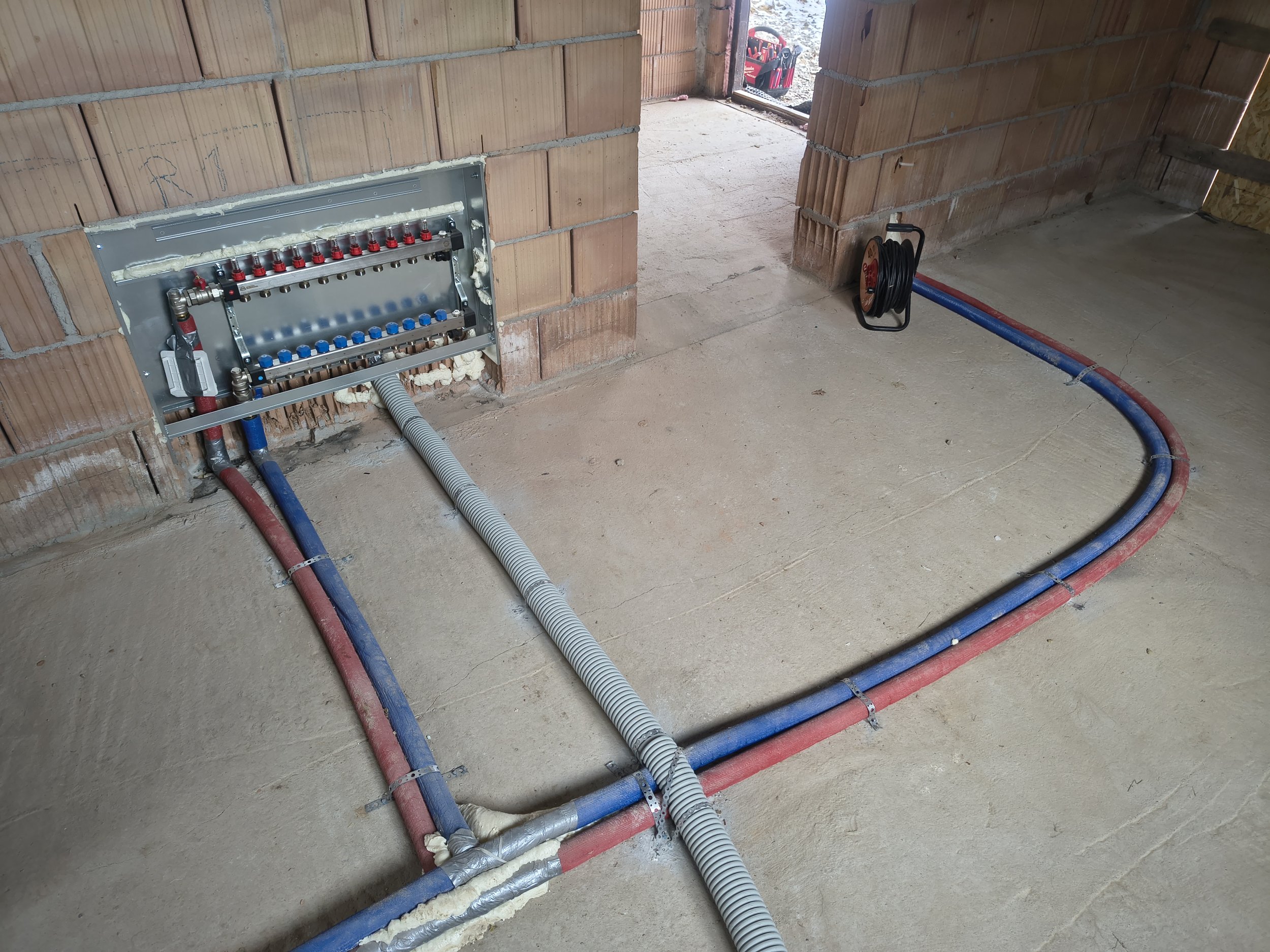 Construction site showing plumbing and electrical conduit installation inside a brick wall. Tools and materials are visible outside the doorway.