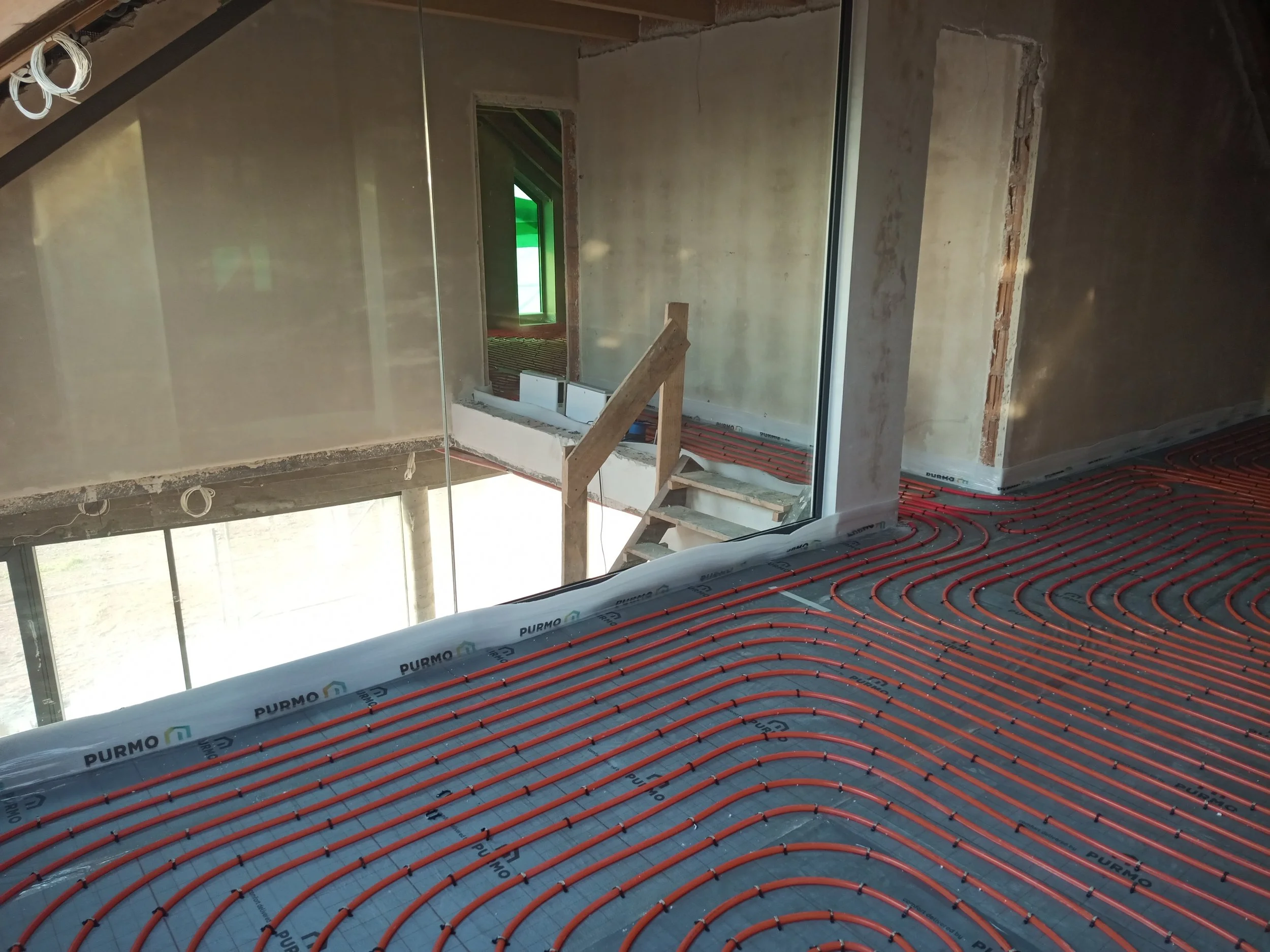 Interior of a building under construction with exposed walls, insulation, a window, and underfloor heating pipes.