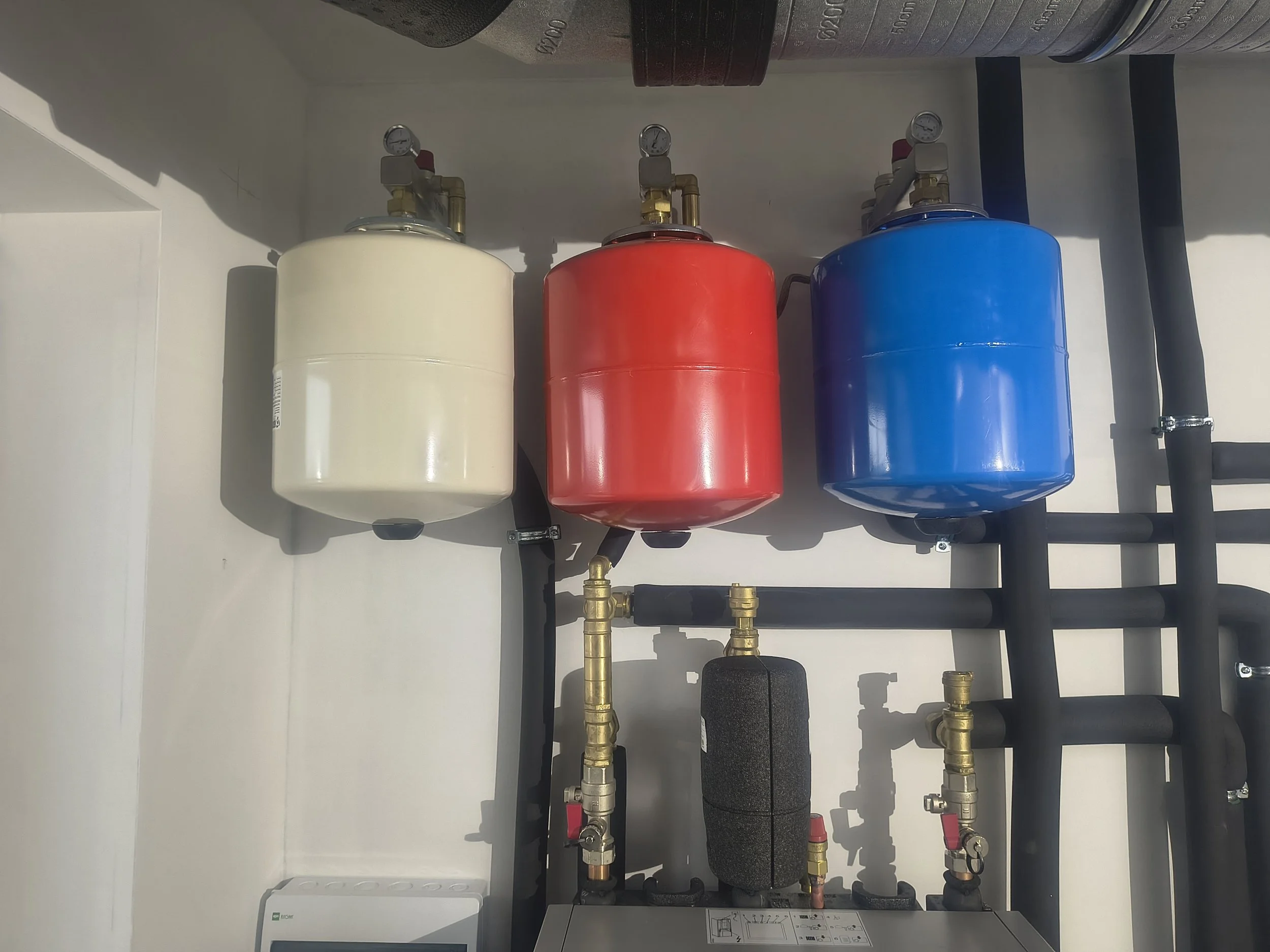 Photograph of three color-coded expansion tanks in white, red, and blue, connected with brass piping and valves, installed under a ceiling in a mechanical or plumbing system.