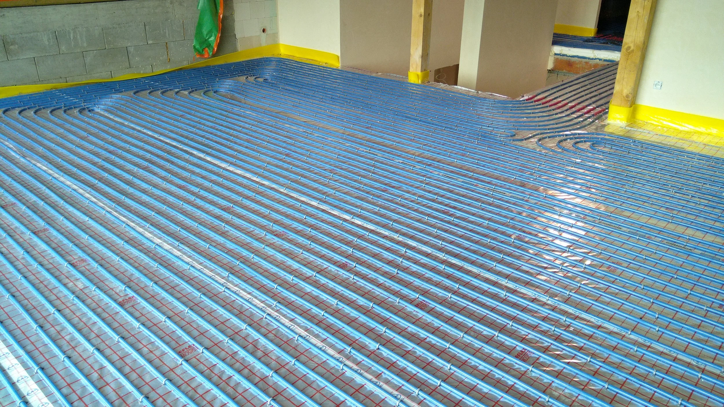 Installation of underfloor radiant heating pipes in a room under construction, with yellow protective barriers along walls.