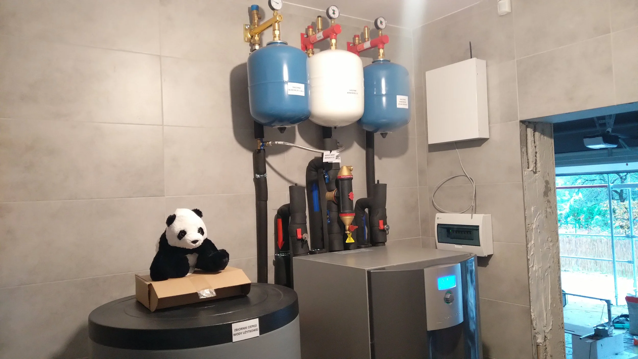 A utility room with blue and white water pressure tanks, piping, gauges, and a control panel. A plush panda toy sits on a cardboard box in the foreground.
