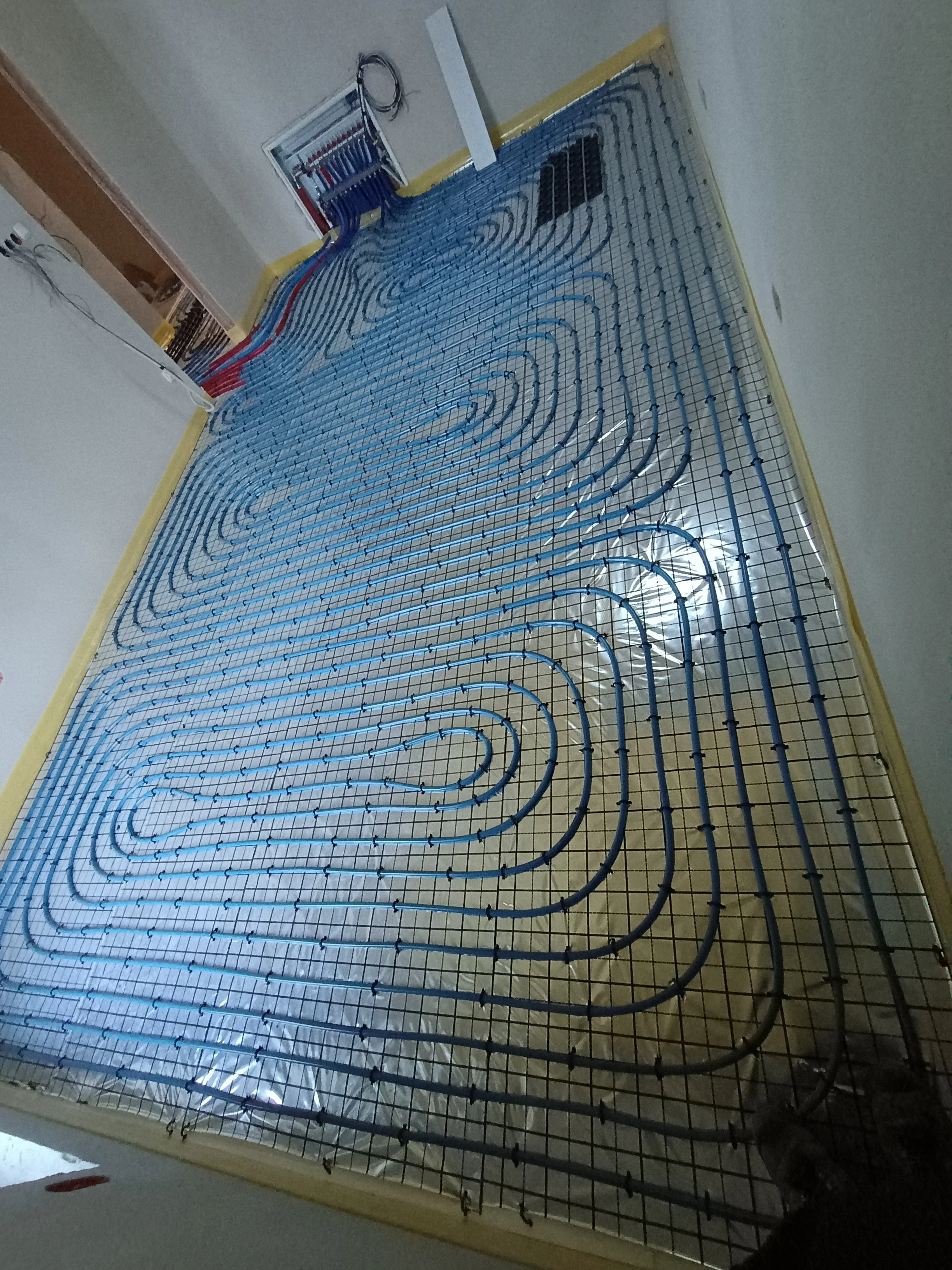 Installation of a radiant floor heating system with blue PEX tubing arranged in a serpentine pattern on a metal grid, in a room under construction.