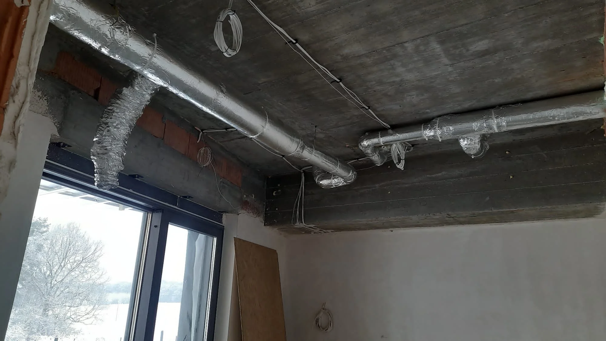 Construction site interior with ductwork and wiring exposed on a wooden ceiling near a large window showing trees outside.