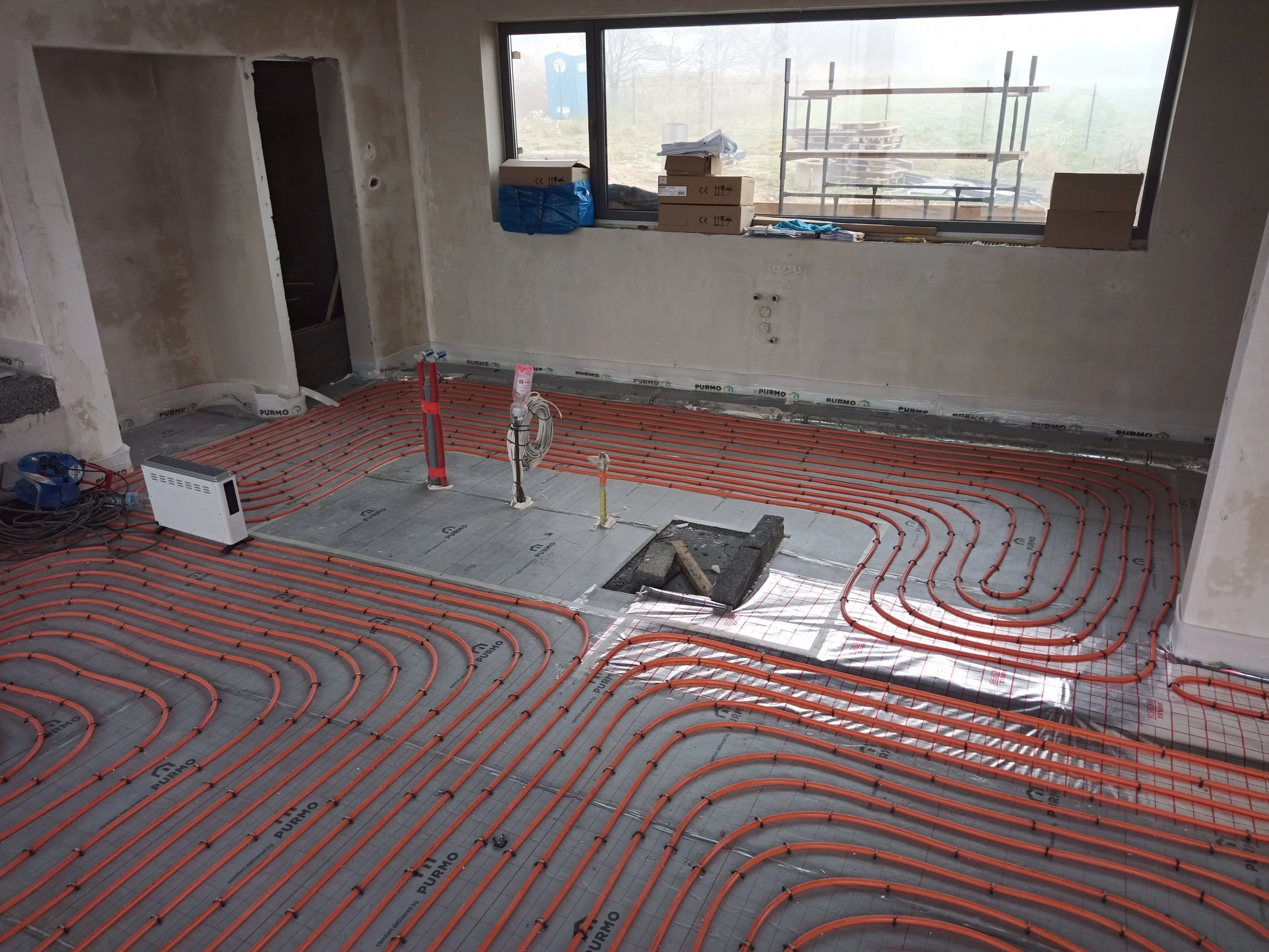 Interior of a room under construction with underfloor radiant heating pipes installed. The pipes are orange and arranged in parallel loops across the floor. There are building supplies and tools on the window ledge and a cardboard box on the window s