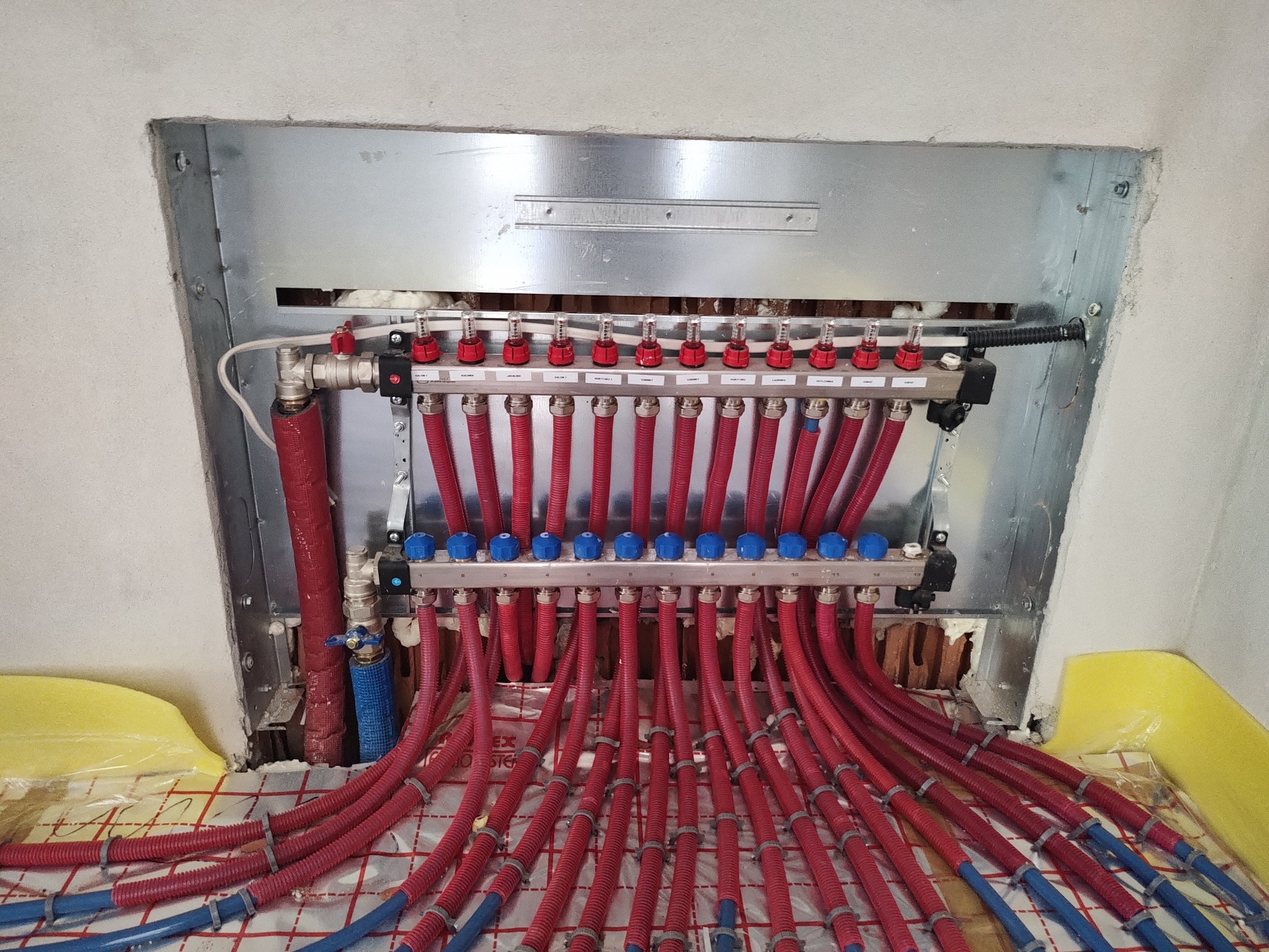 A plumbing installation with red and blue flexible pipes connected to a metallic manifold inside an unfinished wall opening.