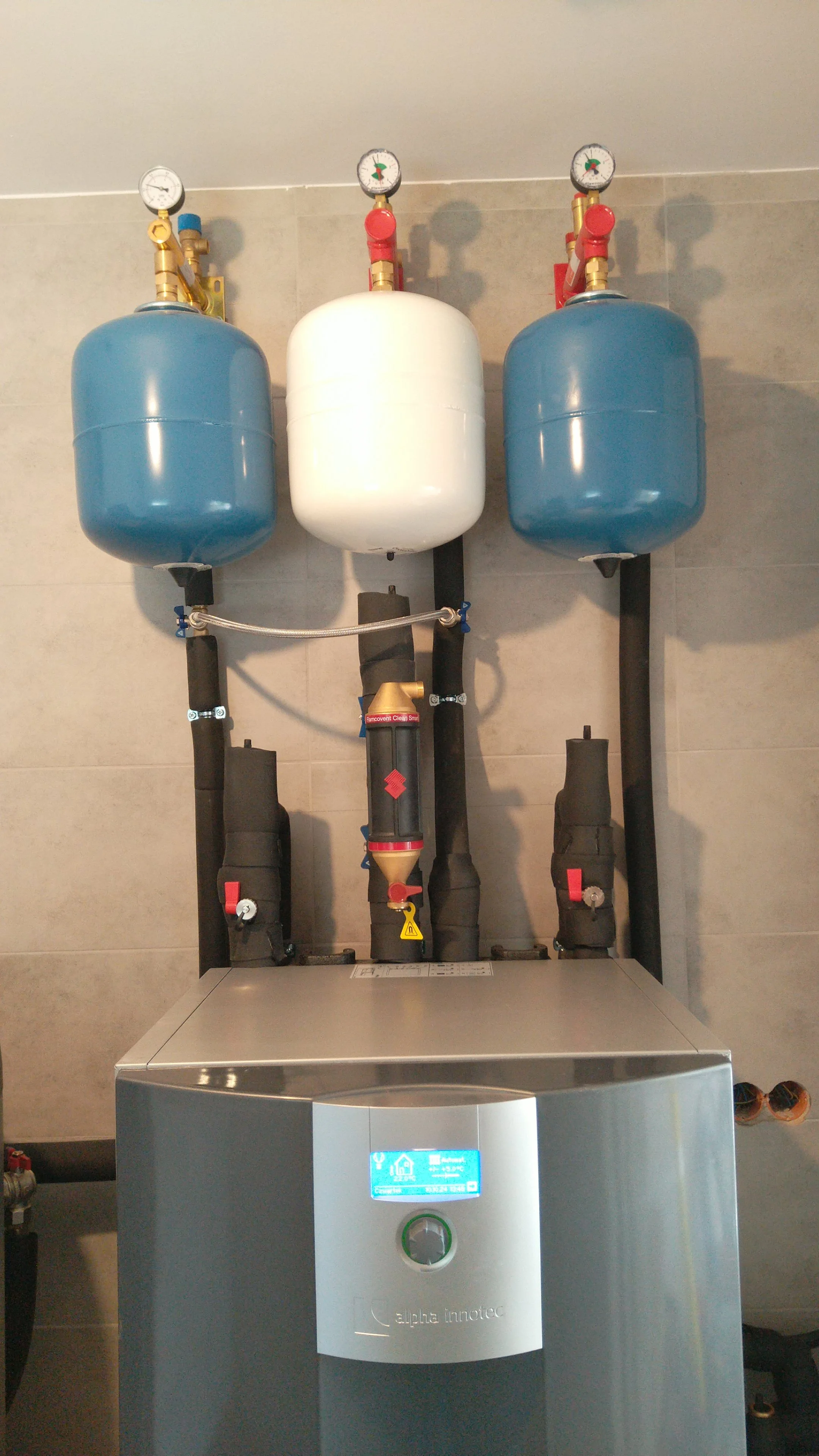 A modern HVAC heating and cooling system with three pressure tanks and gauges mounted on a wall, connected to pipes and control unit.