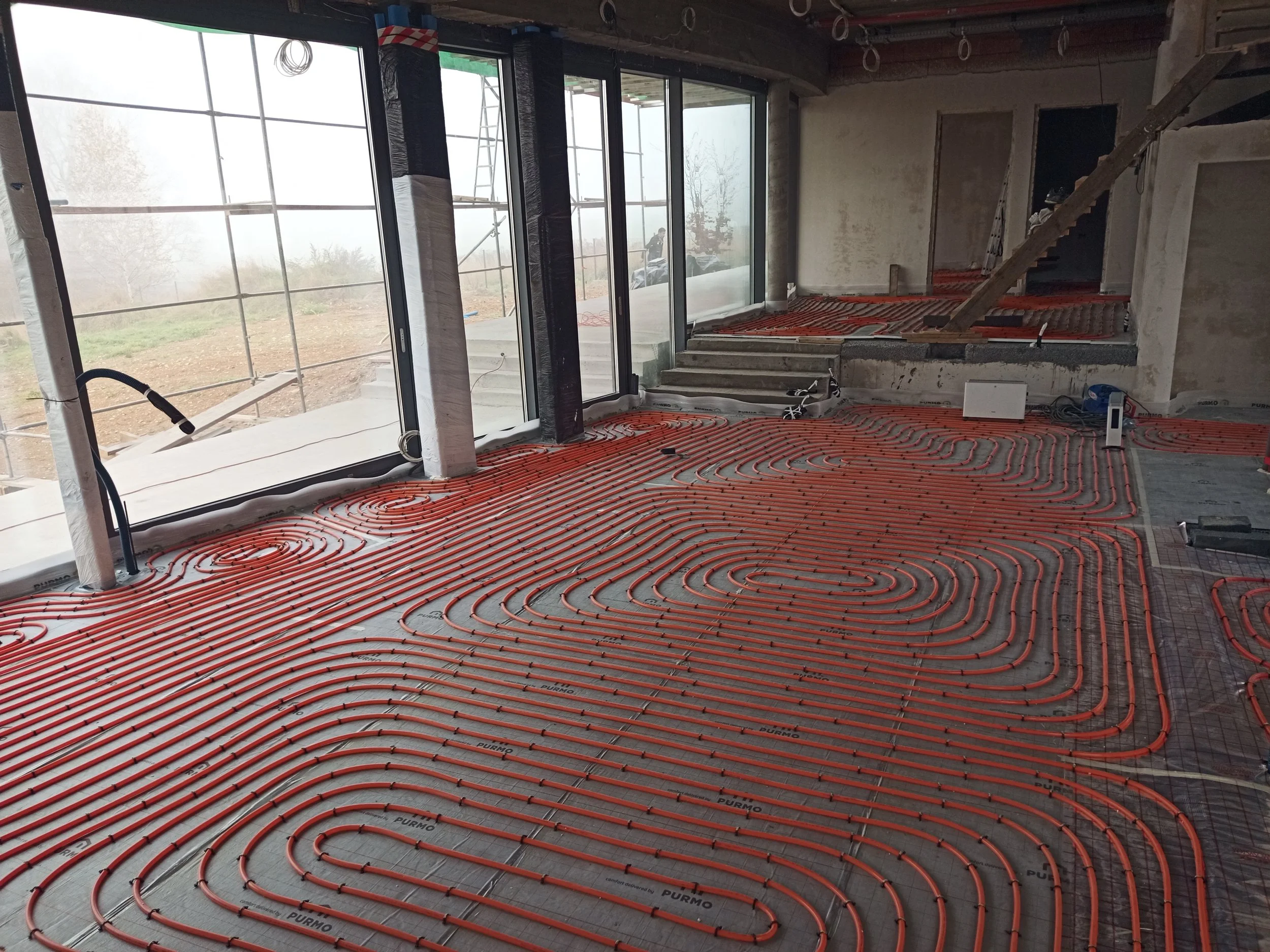 Interior of a building under construction with radiant floor heating pipes installed on the floor. Large floor-to-ceiling windows are visible, showing a foggy outdoor scene with some construction equipment and outdoor scaffolding.