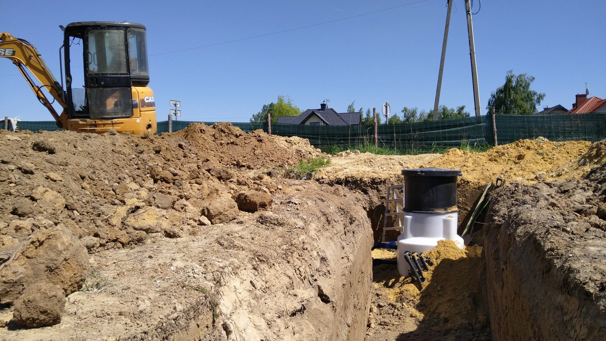Construction site with excavator and open trench, construction materials, and nearby houses under clear blue sky.