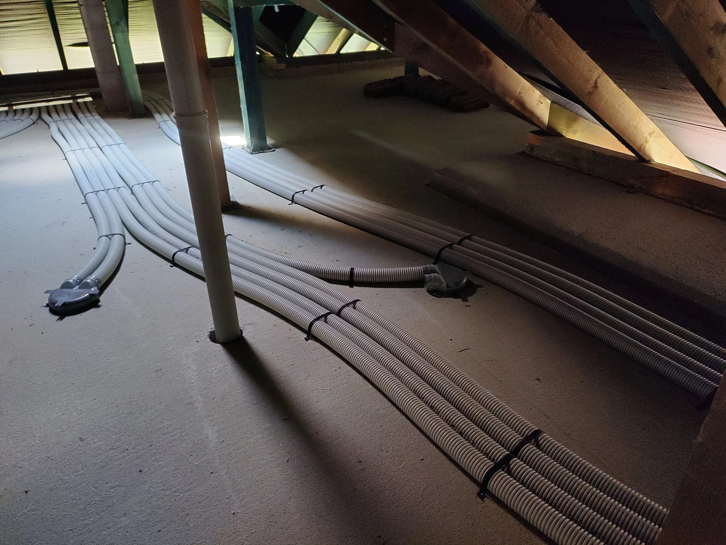 Electrical conduits and cables installed on an unfinished attic floor with exposed wooden beams and insulation.