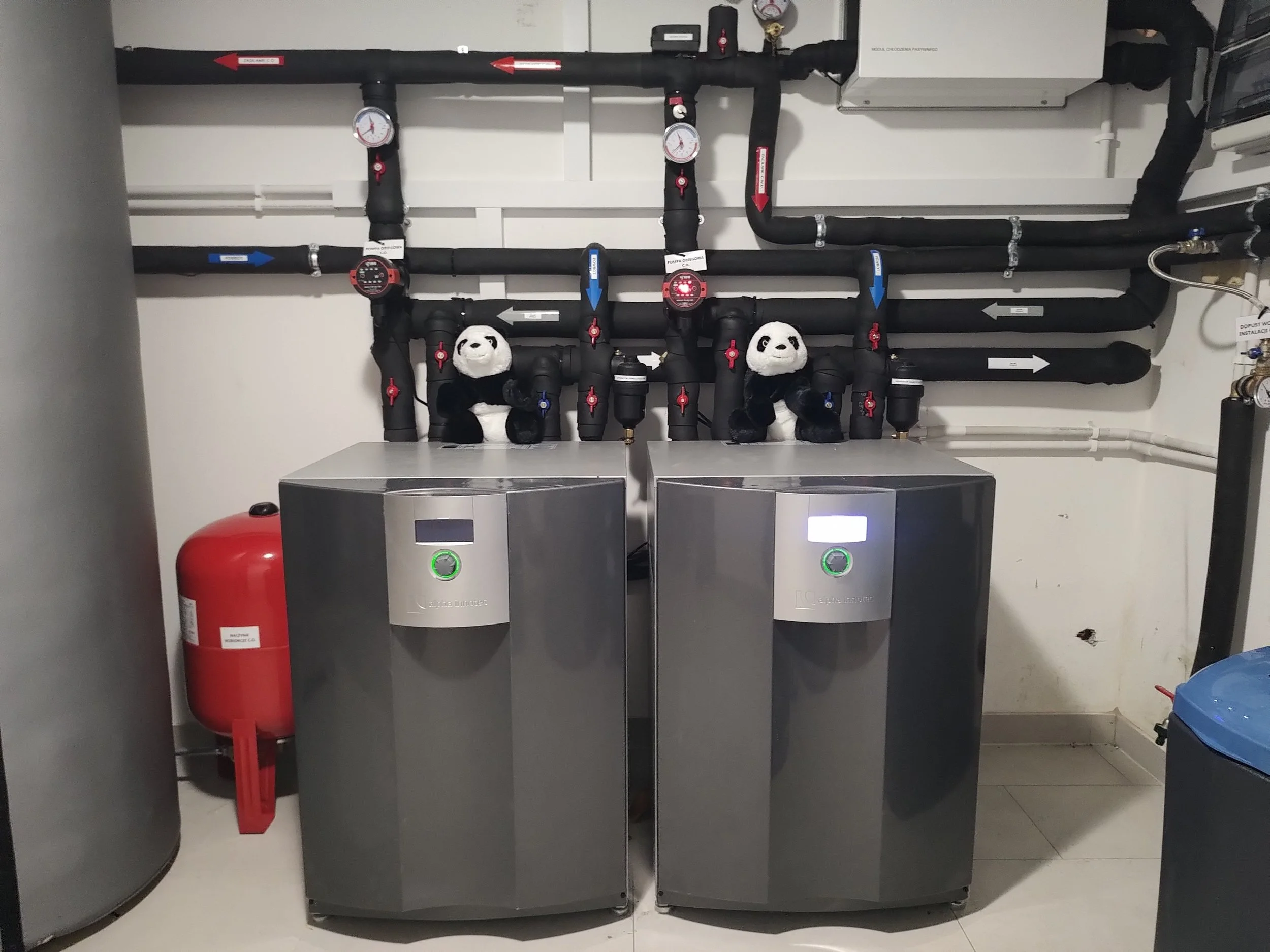 A mechanical room with black pipes, gauges, and control devices mounted on the wall. There are two grey metal units with digital displays, a red fire extinguisher at the side, and plush panda toys placed among the pipes.