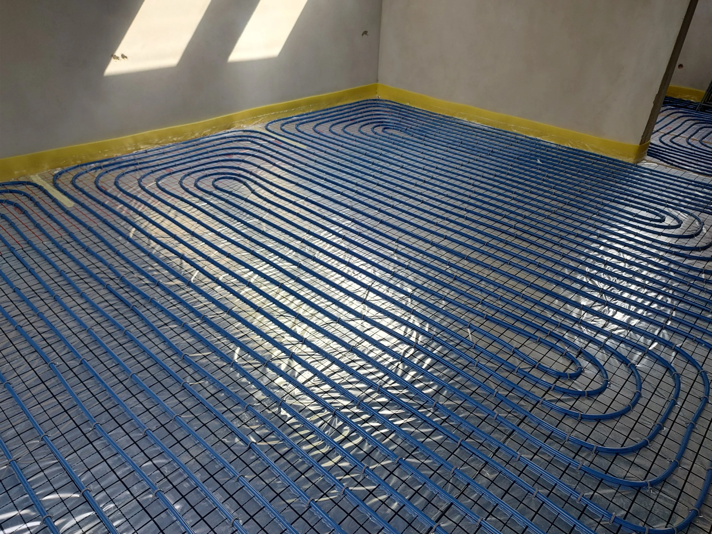 A room with a floor heating system, featuring blue plastic pipes arranged in a serpentine pattern on top of a metal mesh. The walls are plain and light-colored, with sunlight coming through a window and casting shadows on the wall.