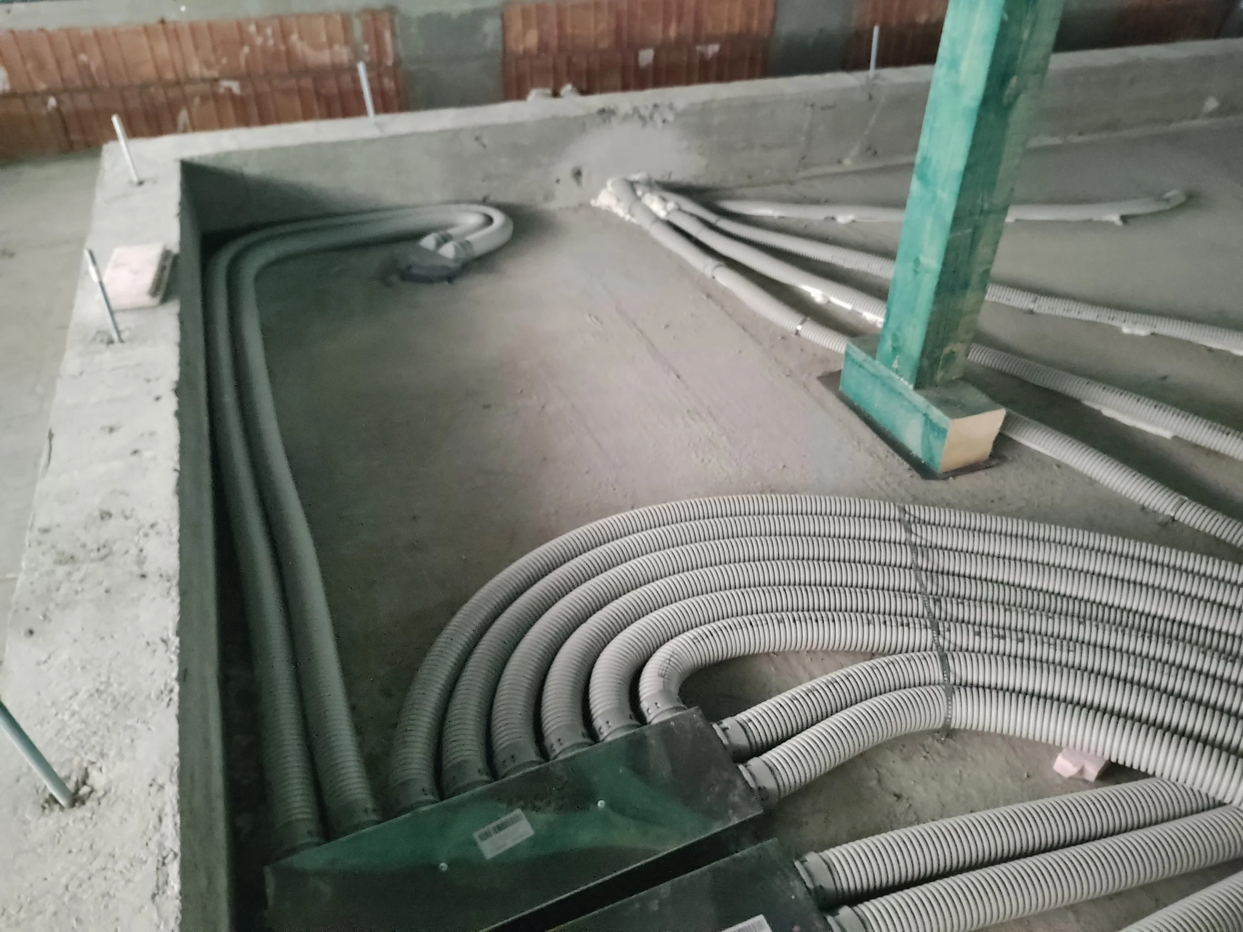 Construction site with a concrete wall, green wooden support post, and multiple white flexible electrical conduit pipes on the floor.