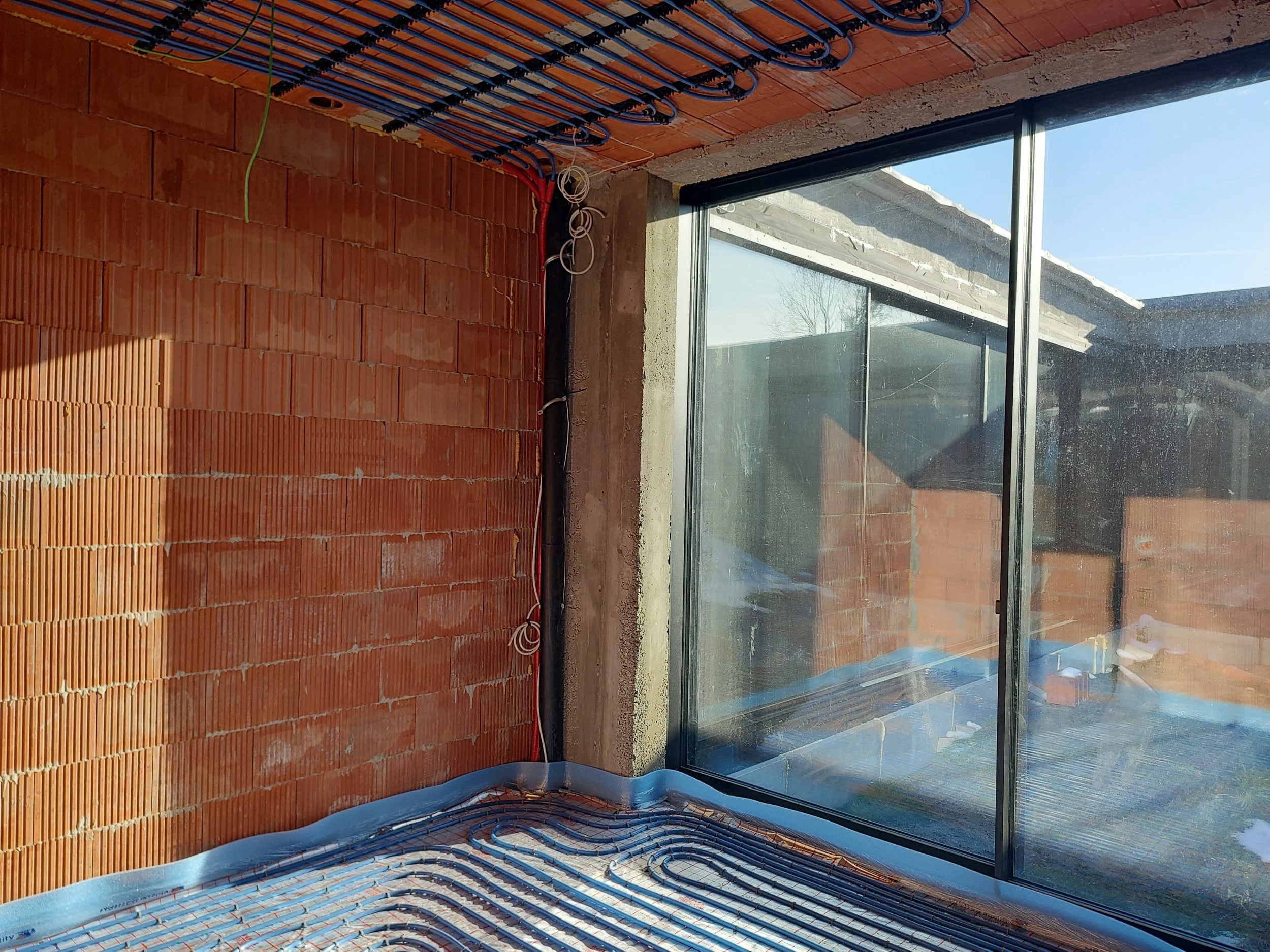Interior of a building under construction with exposed red brick walls, electrical wiring, and a large window showing an outdoor patio with a roof, blue sky, and trees.