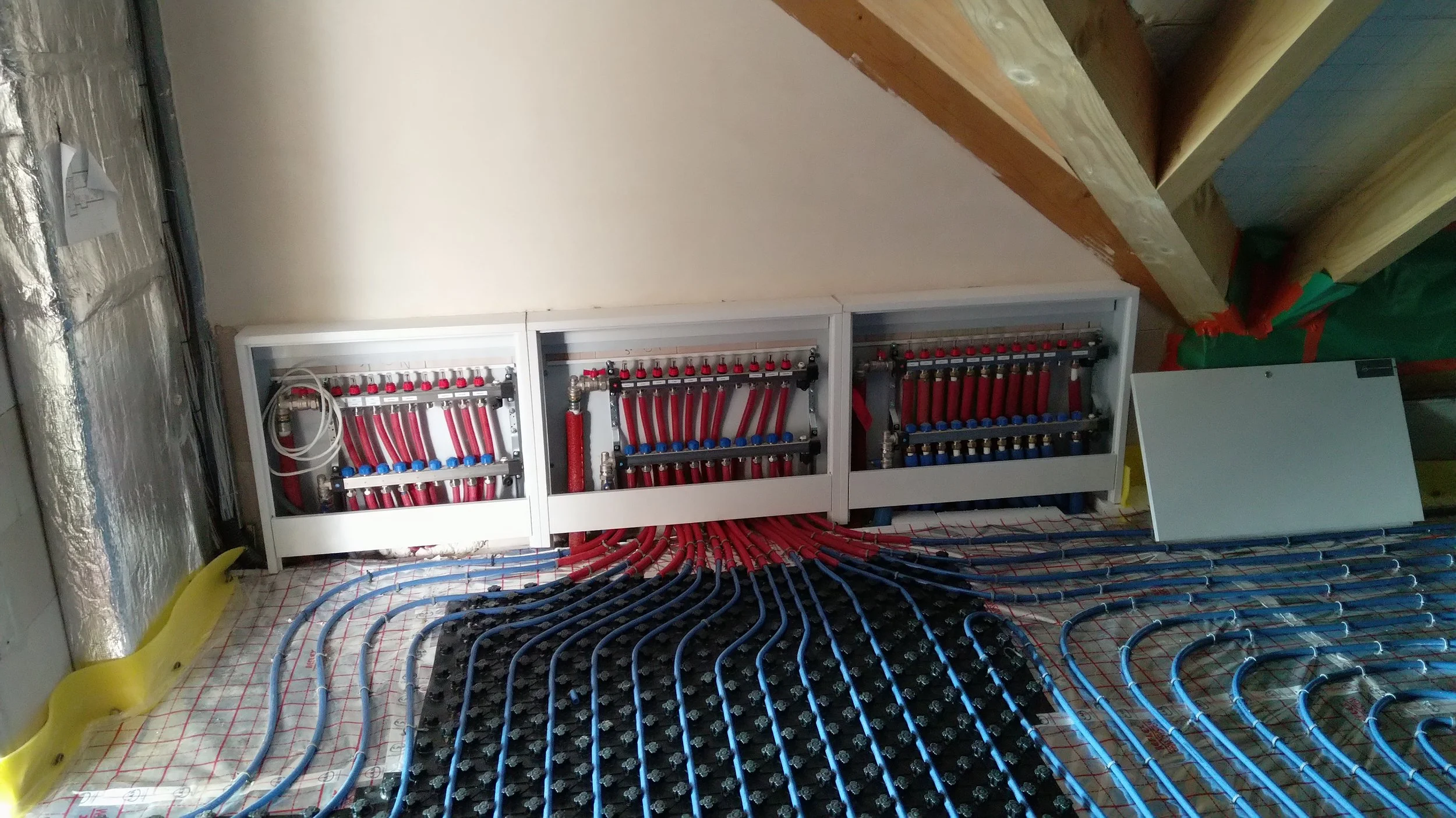 A partially constructed electrical or plumbing control panel with multiple red and blue pipes, black mounting plates, and white framing in a room with a wooden beam ceiling and reflective insulation on the walls.