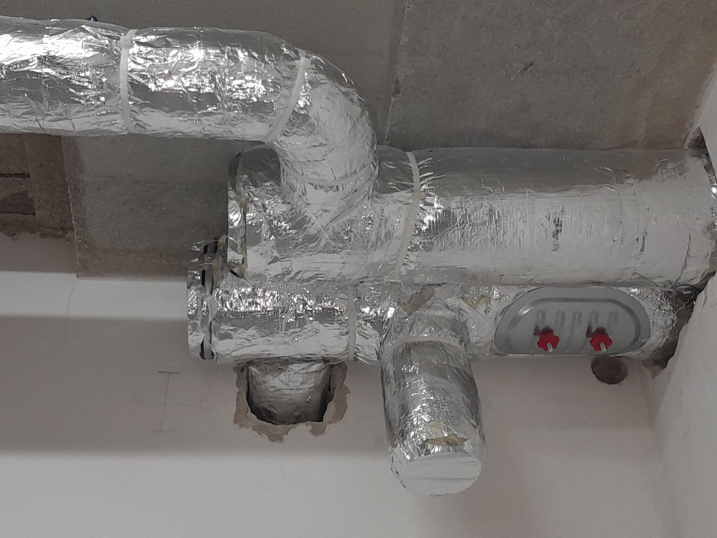 A series of HVAC duct pipes wrapped in silver foil insulation, connected in a complex network on a wall.