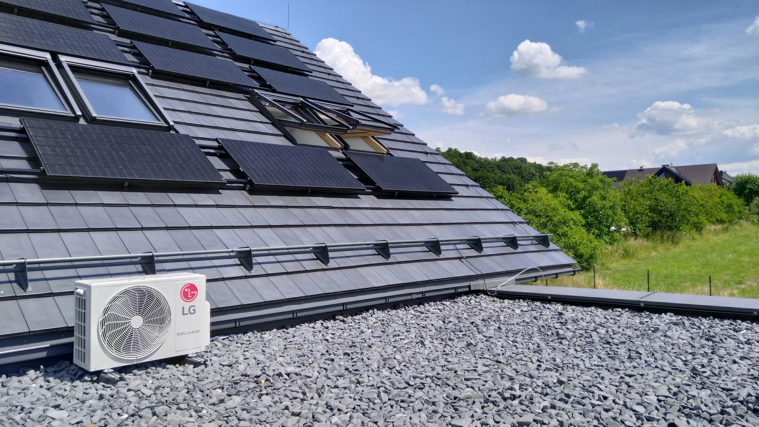 Residential rooftop with multiple solar panels installed, an air conditioning unit at the bottom, and a gravel surface on the roof. Houses and green trees are visible in the background under a blue sky with clouds.