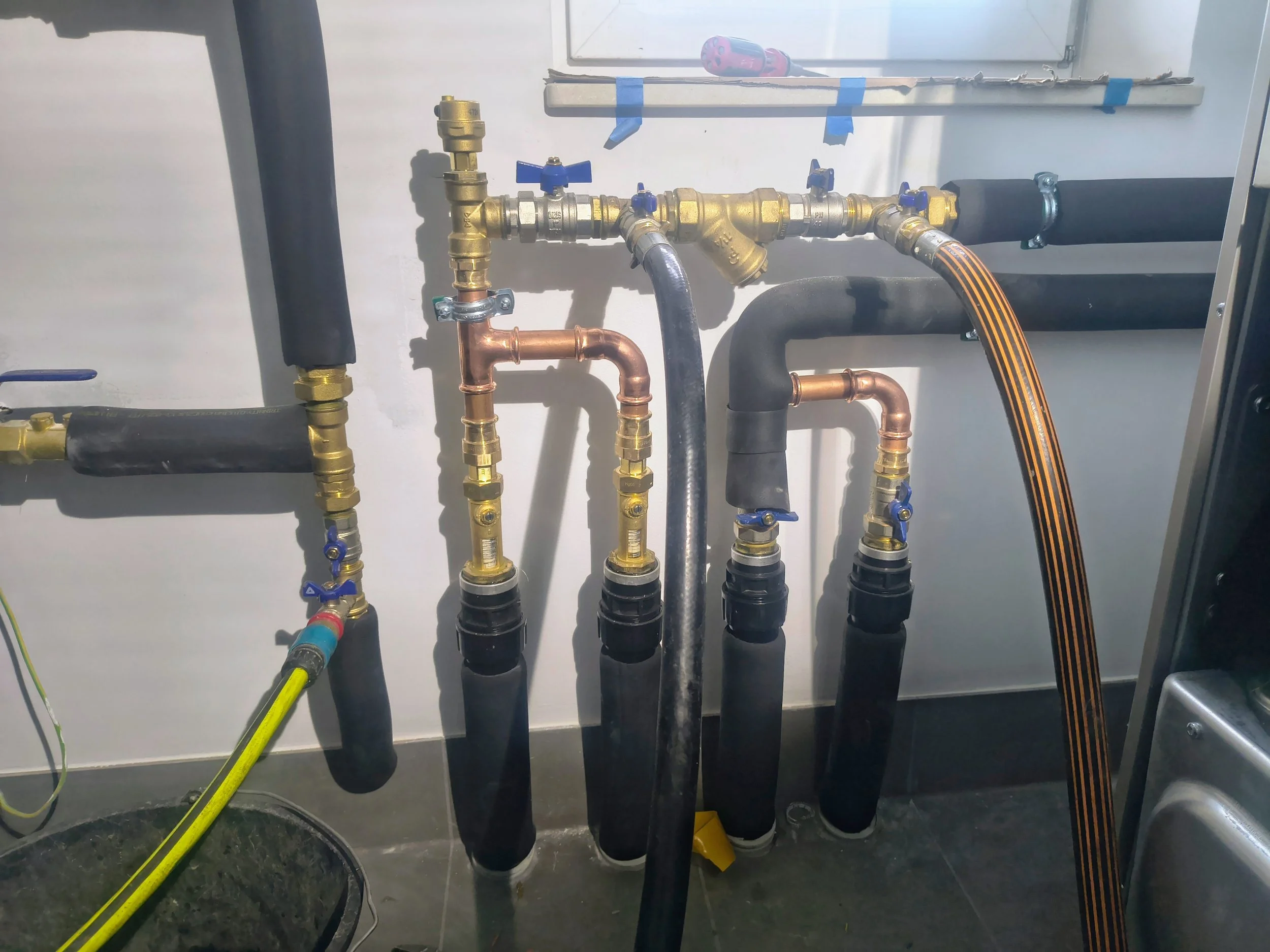 A series of plumbing pipes and valves connected to a wall, including black, copper, and brass components, with some blue handles and insulation, in a utility or mechanical room.