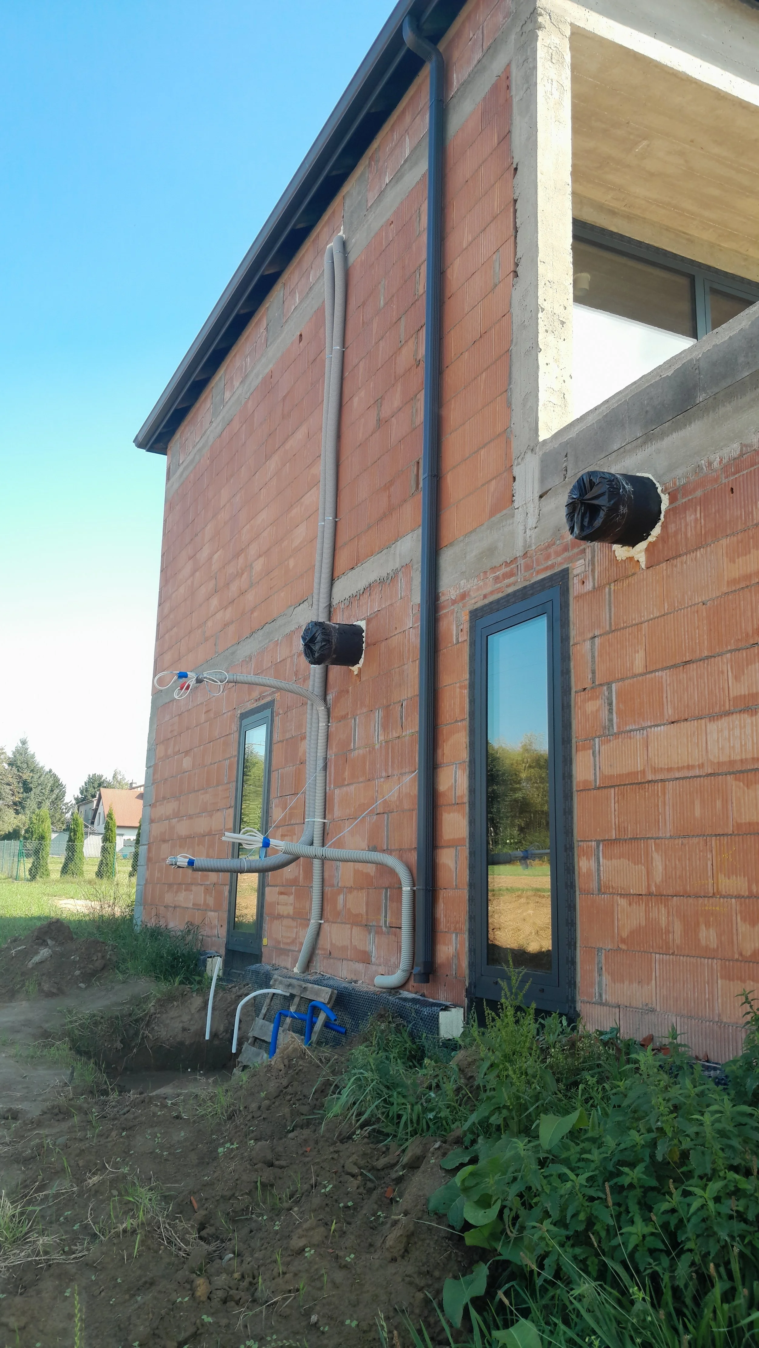 Construction site of a residential building with exposed brick walls, electrical conduits and plumbing pipes installed. The building has large windows and the area around it has dirt and green plants.