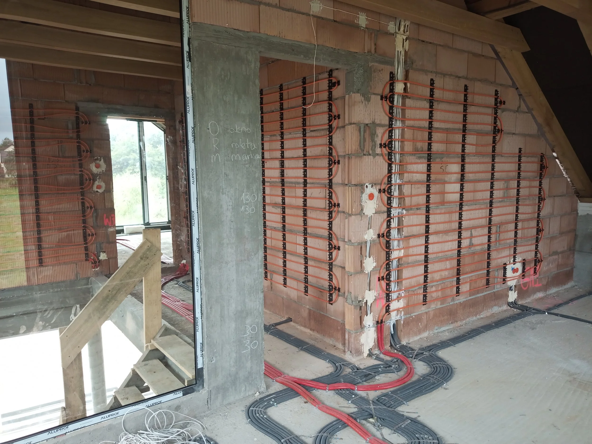 Interior view of a building under construction showing exposed brick walls, electrical wiring, and radiant floor heating cables installed on the floor. A concrete wall with handwritten measurements in white chalk is also visible.