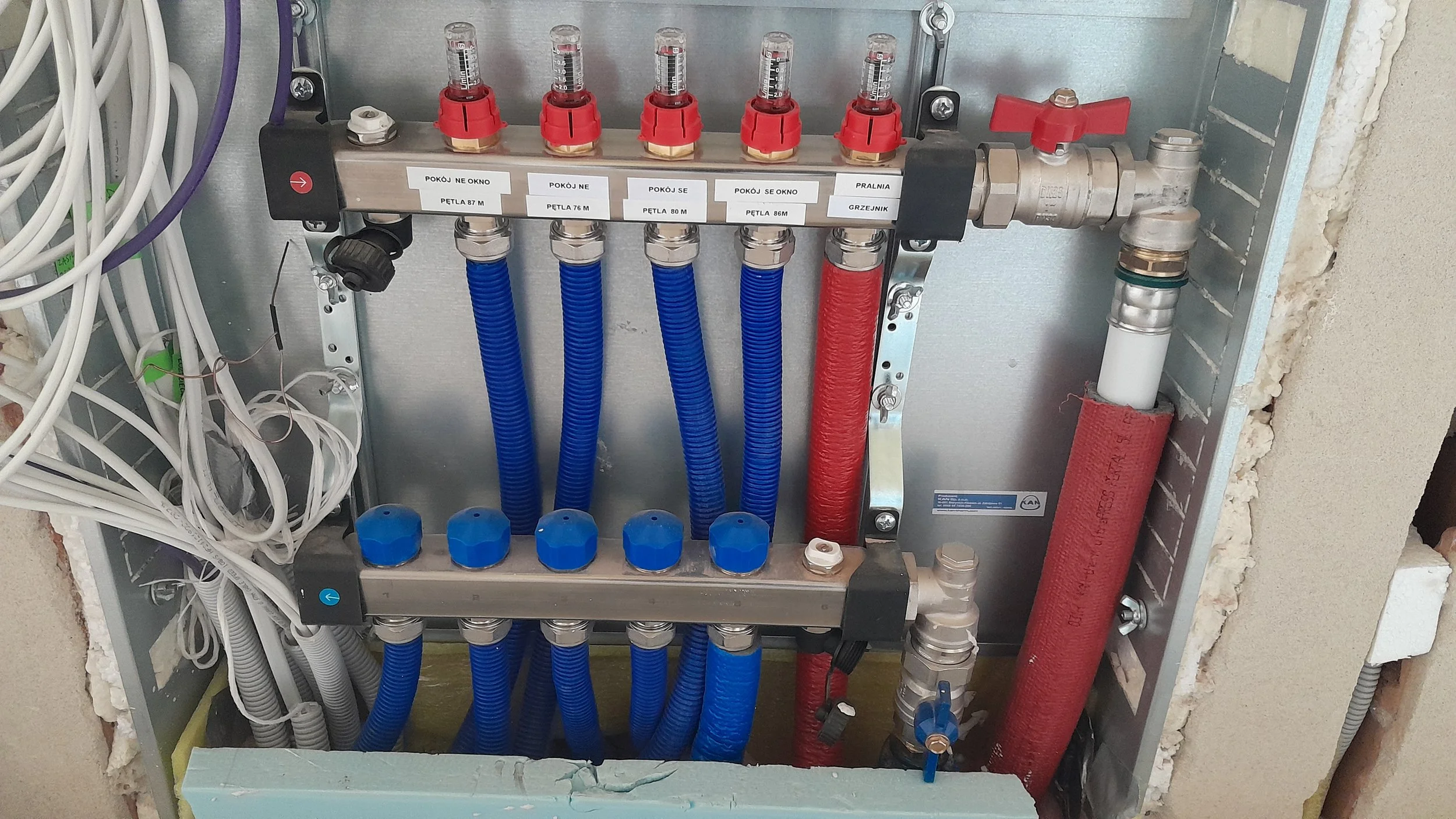 A plumbing manifold with blue and red flexible hoses connected, labeled with different room names and circuit numbers, installed in a wall with electrical wires nearby.