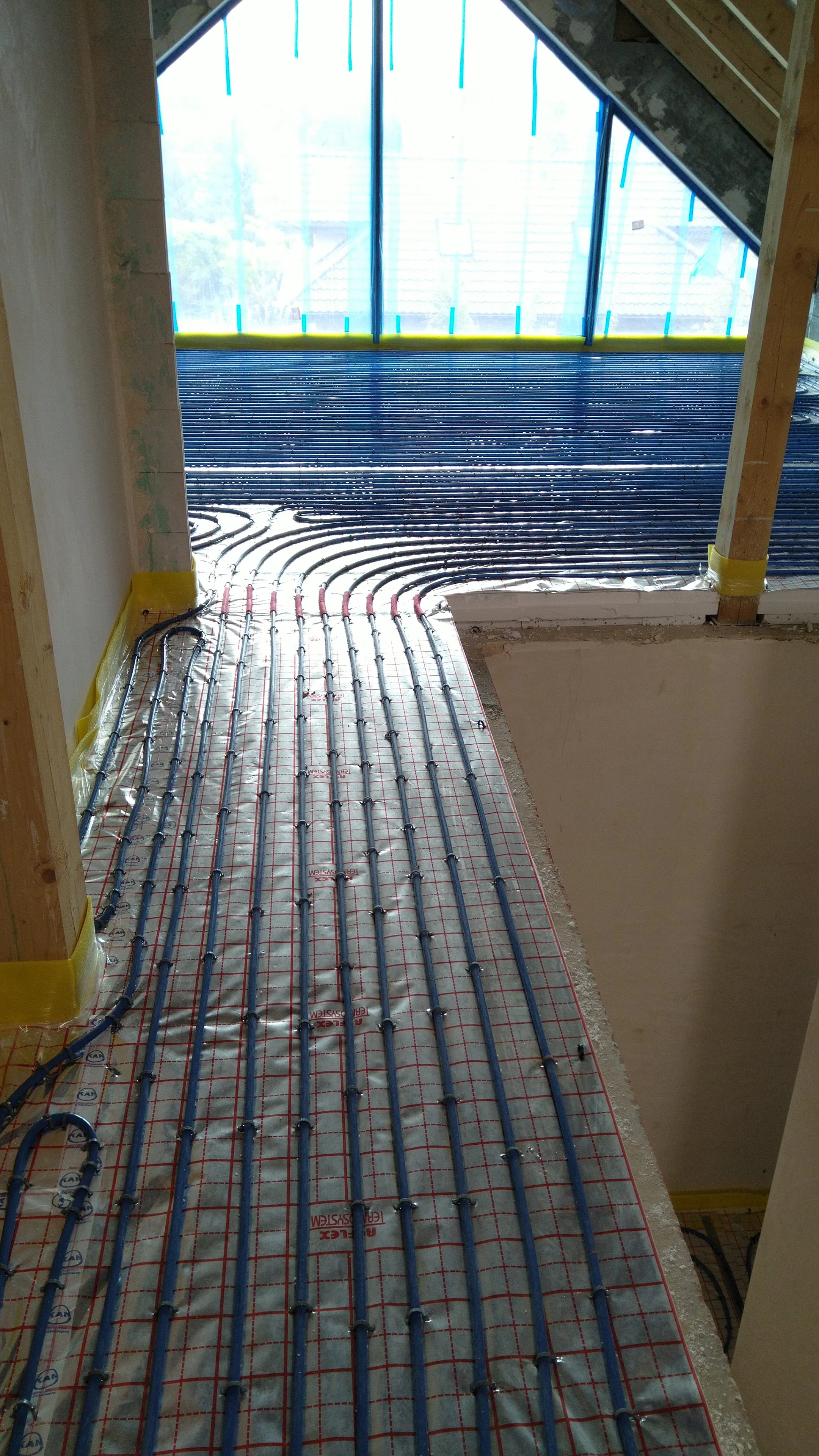 Underfloor heating pipes being installed in a room with large windows, showing black plastic pipes laid on a reflective surface with red grid lines for positioning.
