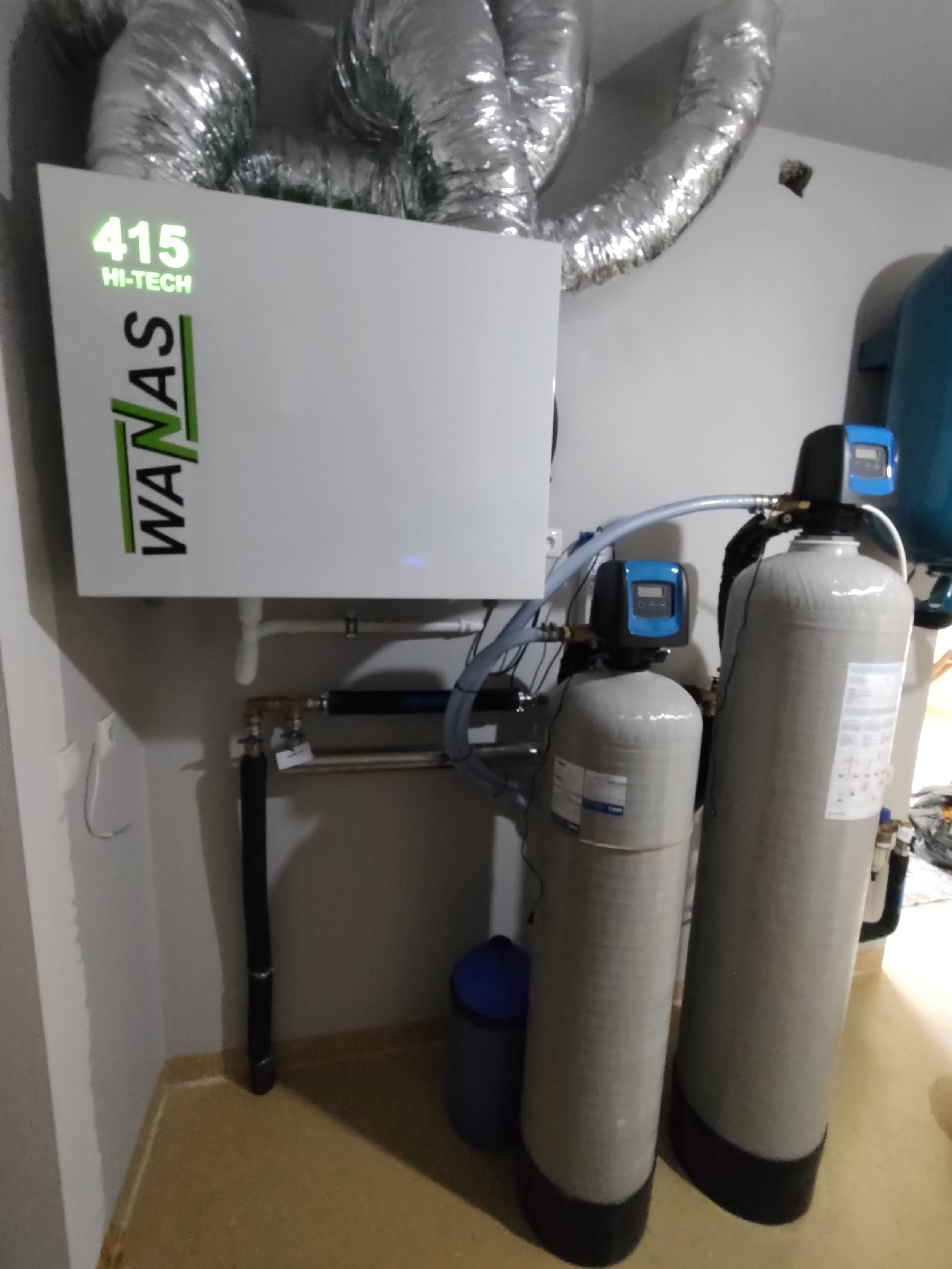 A water softener system with a control panel labeled '415 HI-TECH' and multiple large cylindrical tanks connected with pipes and hoses in a utility room.
