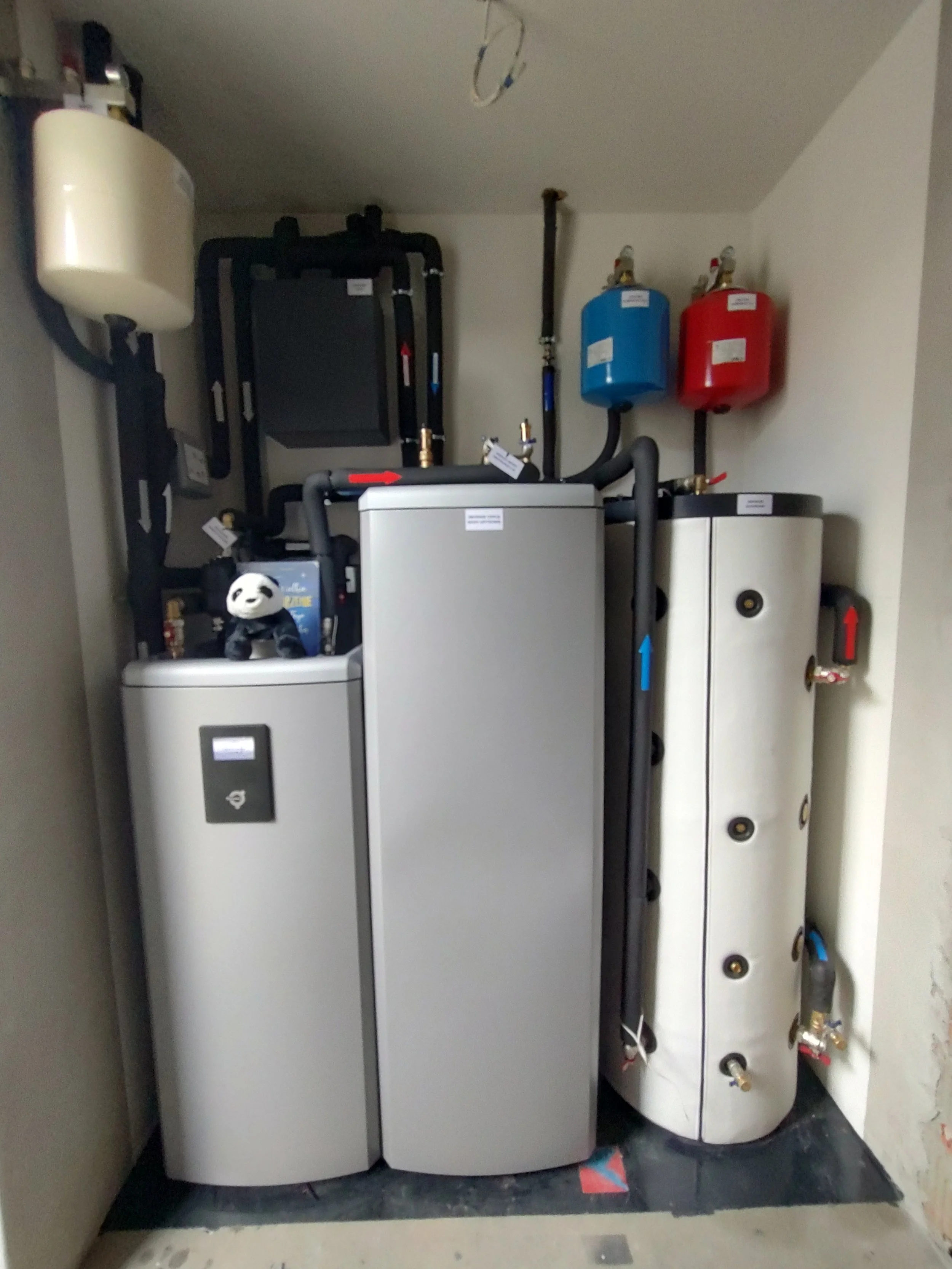 A utility room containing a water heater, a boiler, and two expansion tanks installed on the wall, with pipes connected to each appliance.