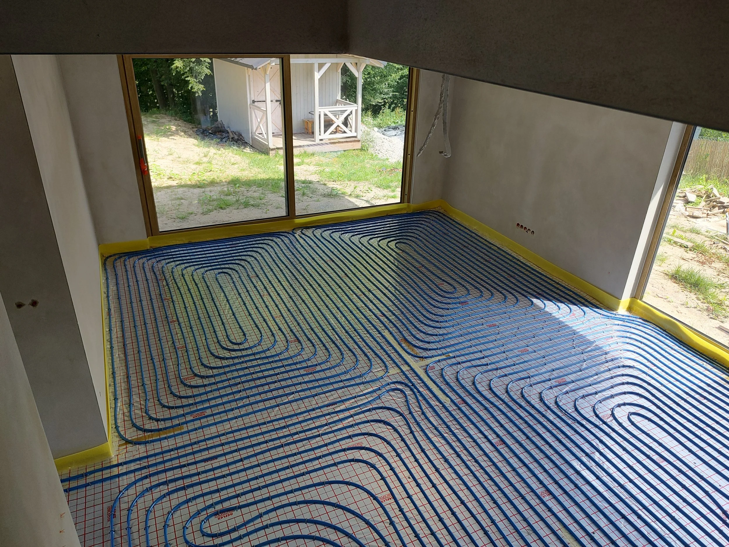 Interior view of a room under construction with radiant floor heating pipes installed over a floor surface, overlooking a backyard with a small shed and green trees outside sliding glass doors.