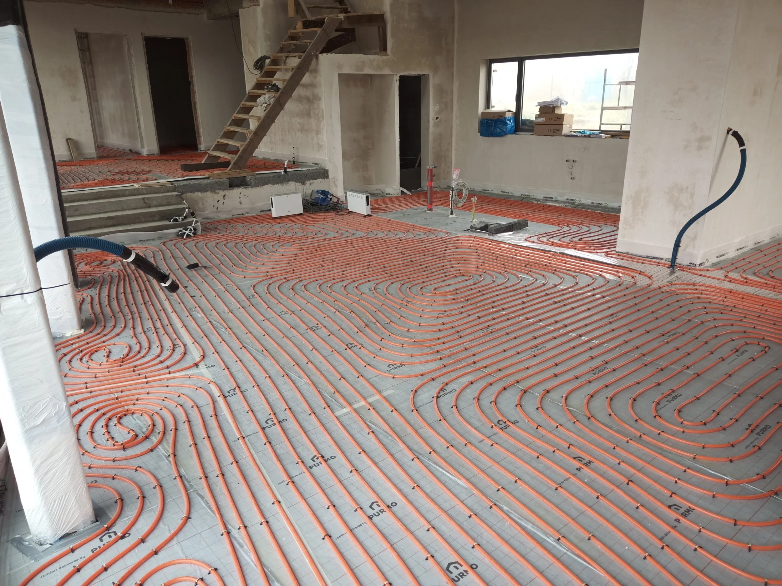 A room under construction with orange radiant floor heating pipes installed over insulation. There are stairs, construction tools, boxes, and unfinished walls in the space.