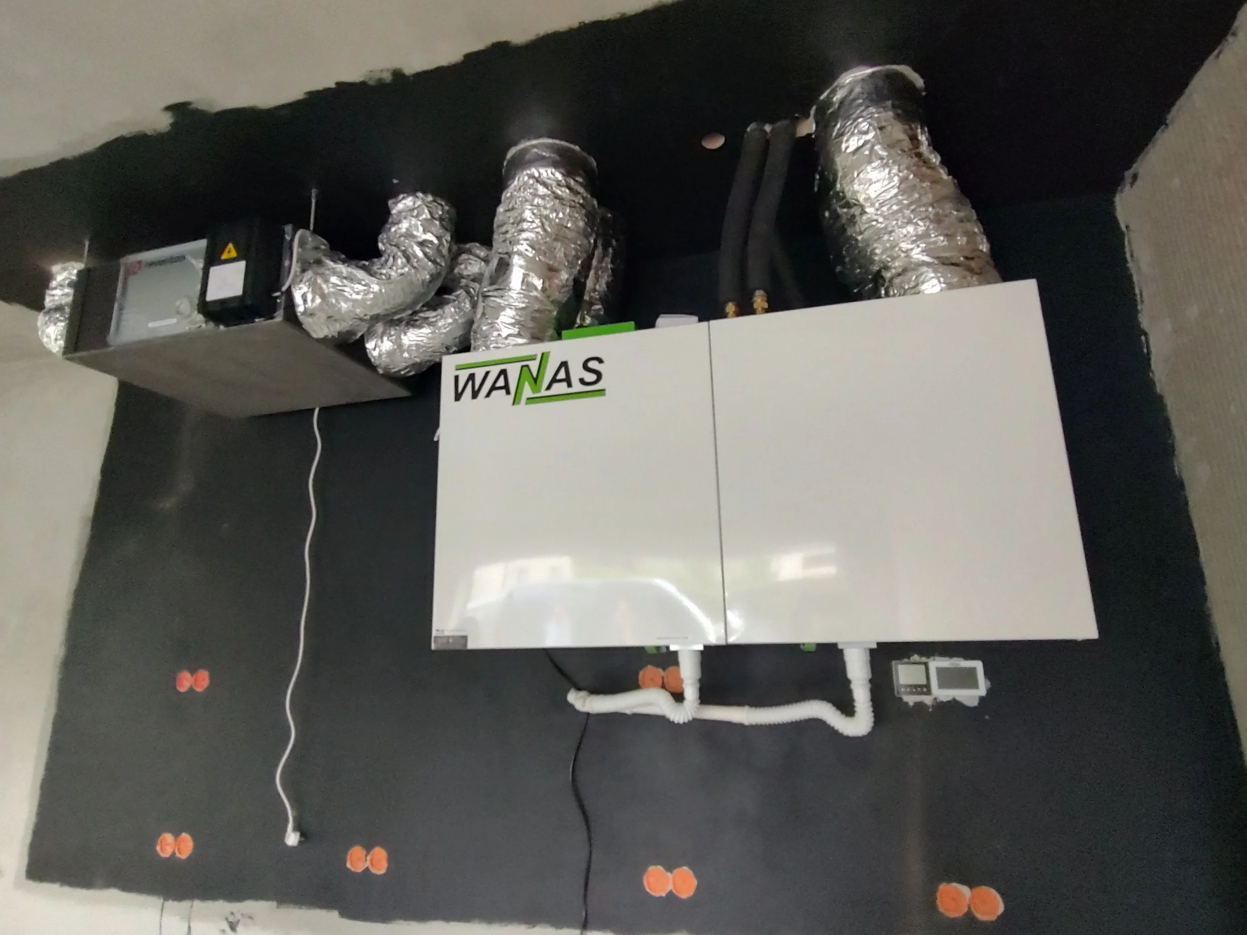 A white box labeled 'WANAS' installed on a black wall with ductwork above and orange circular objects on the floor.
