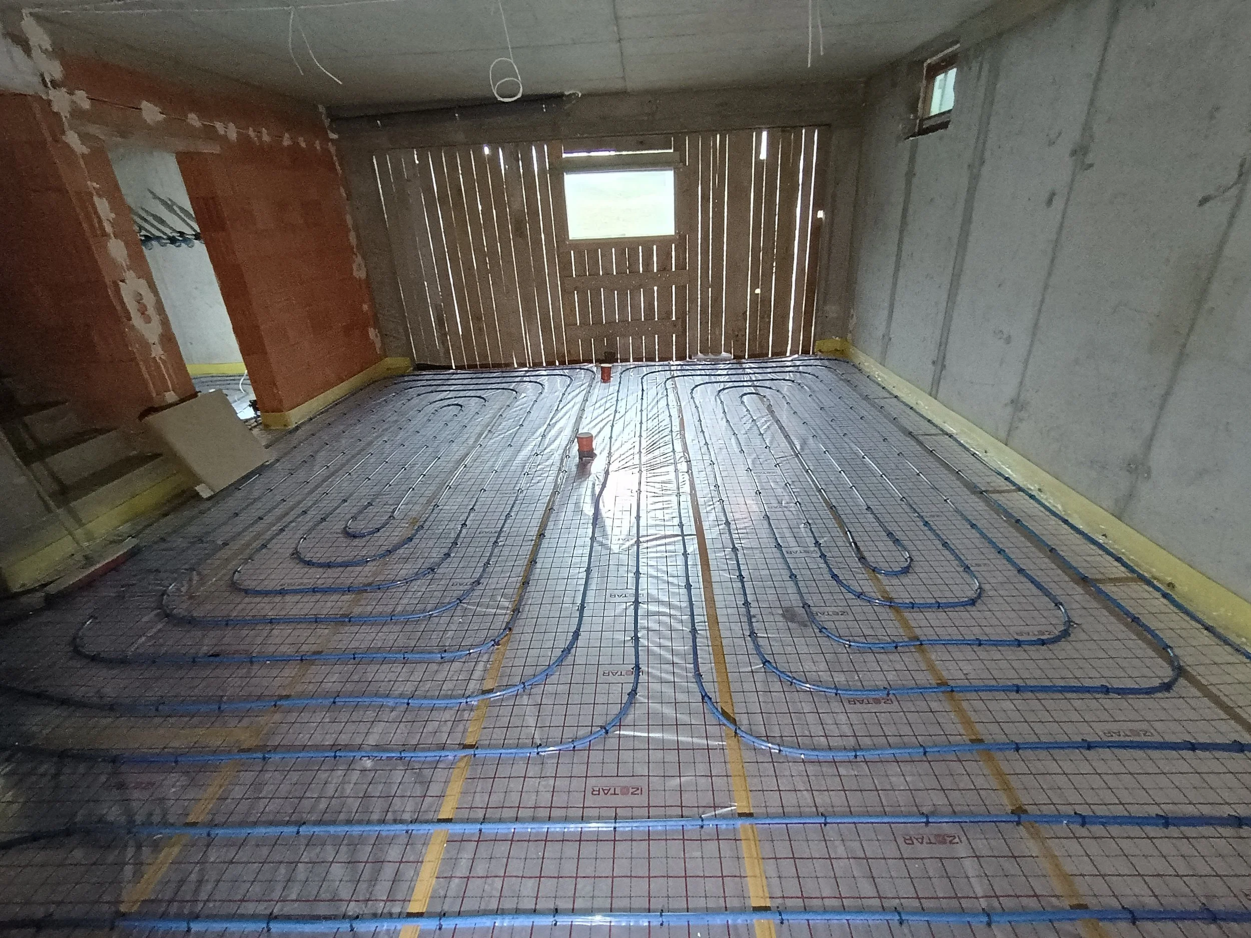 Interior of a room under construction with a radiant floor heating system installed on the floor and wooden slat walls, small windows, unfinished ceiling, and construction materials scattered around.