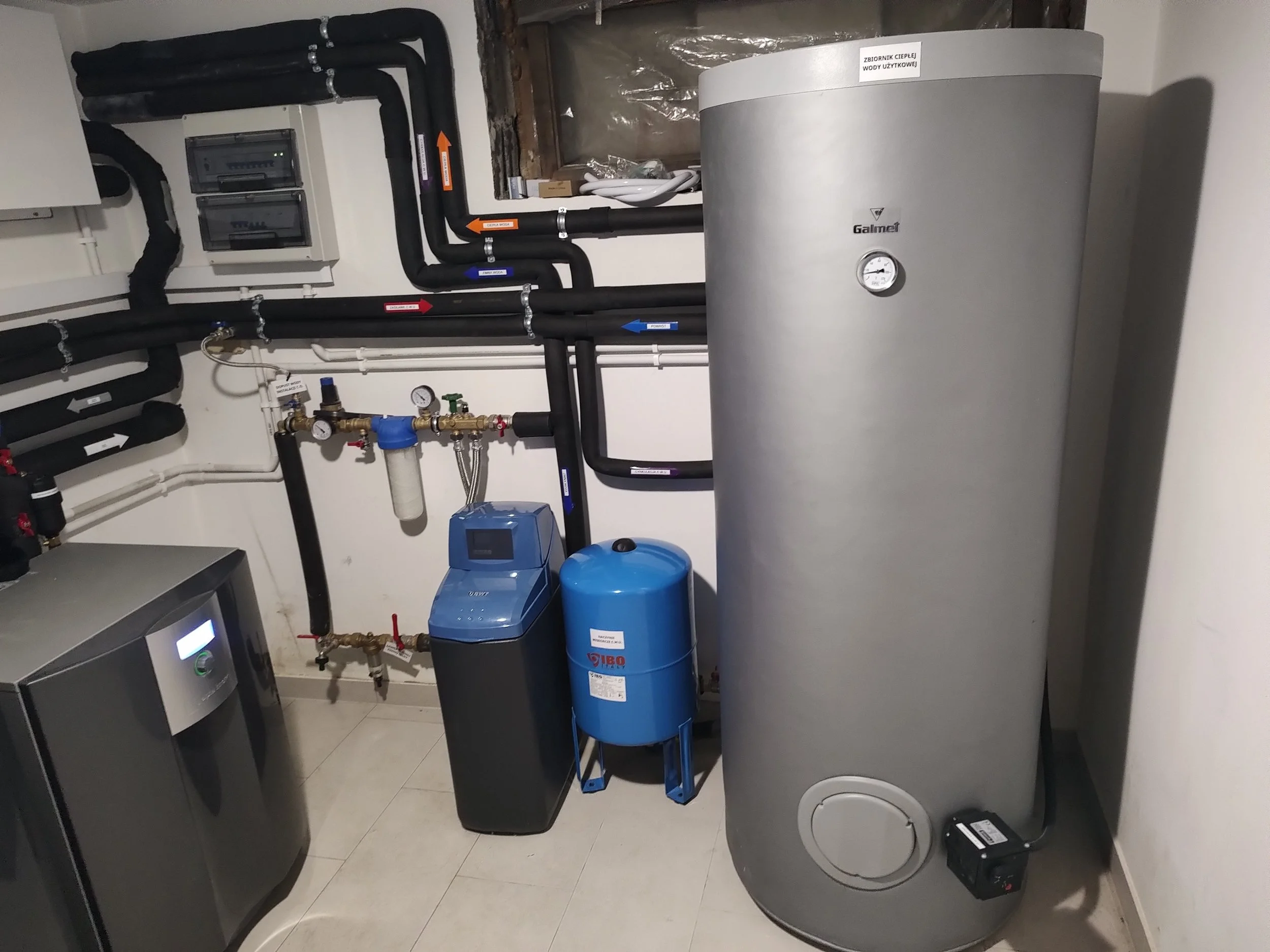 Mechanical room with pipes, gauges, water heater, and filtration system. Large cylindrical water heater on the right labeled 'Galmet,' and a blue water filter in the center.