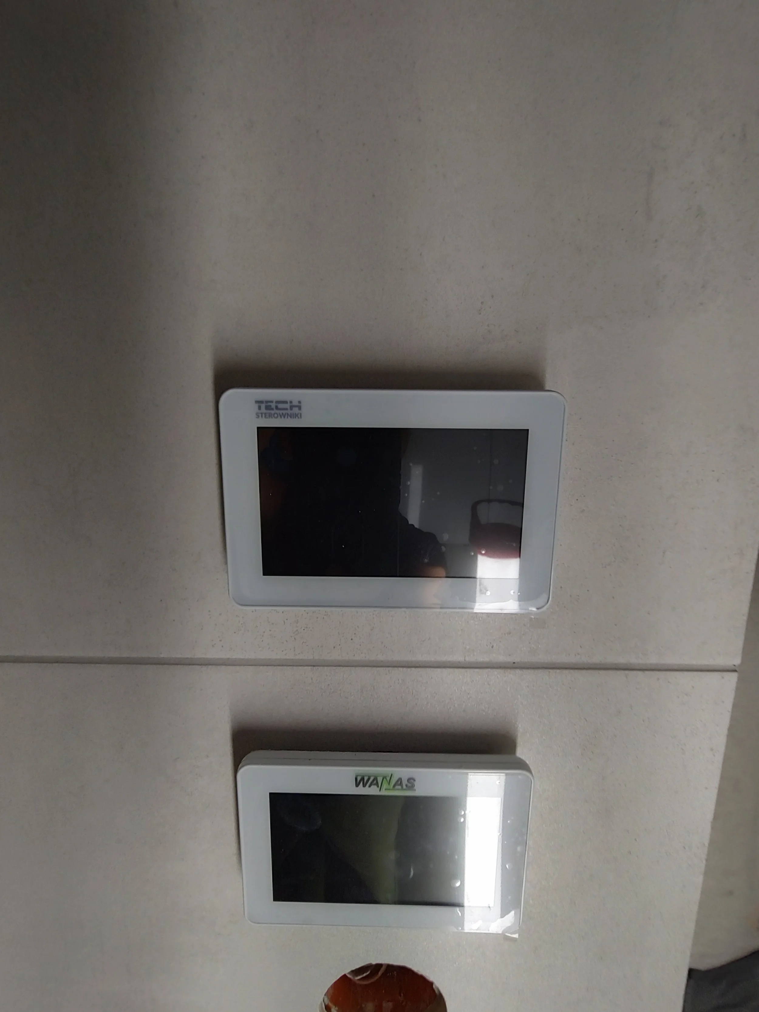 Two white digital devices mounted on a white wall; one with a black screen labeled 'TECH' and the other with a black screen labeled 'WAS'.