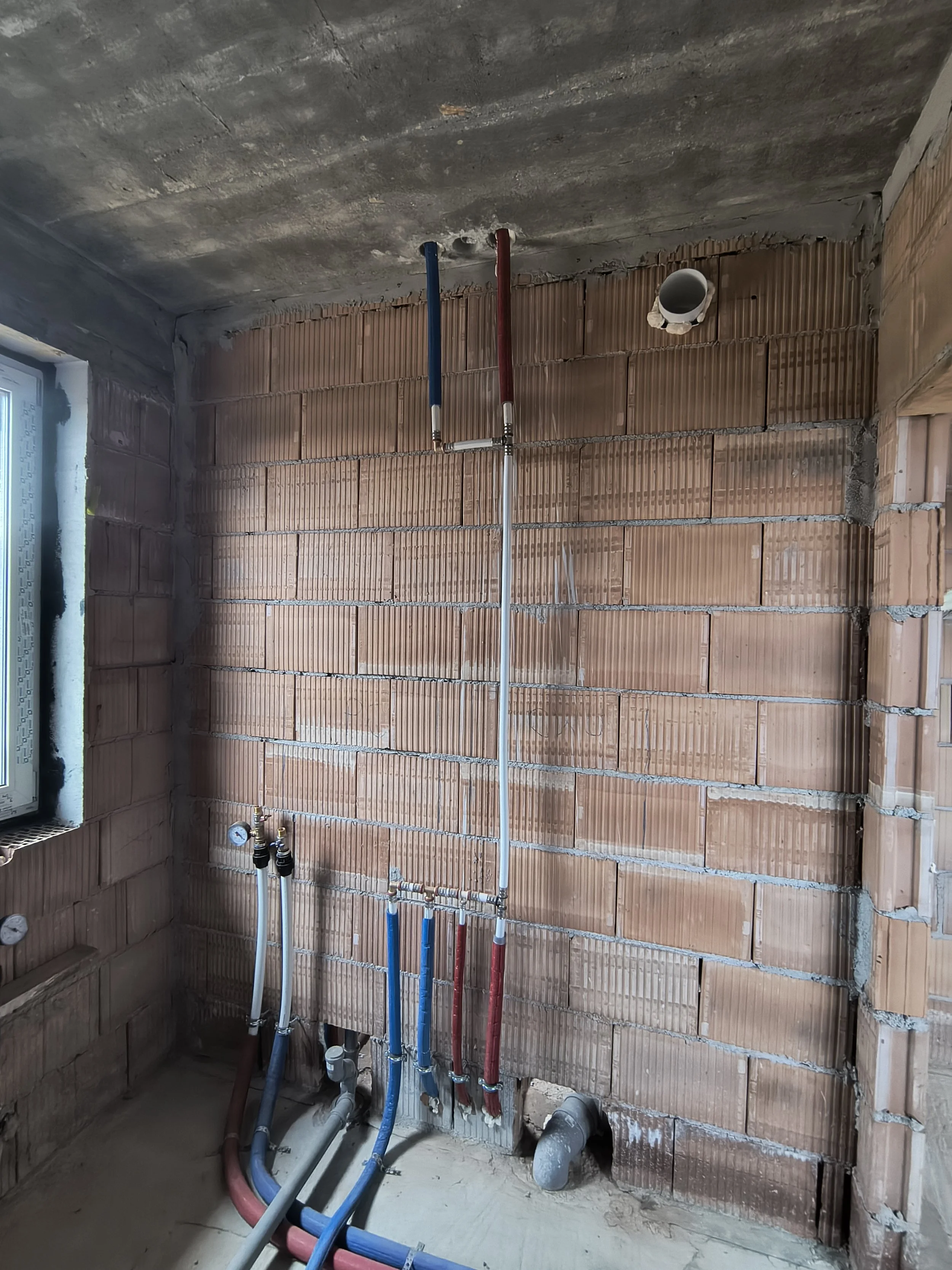 Interior of a building under construction with exposed brick wall and plumbing pipes in red, blue, and gray colors.