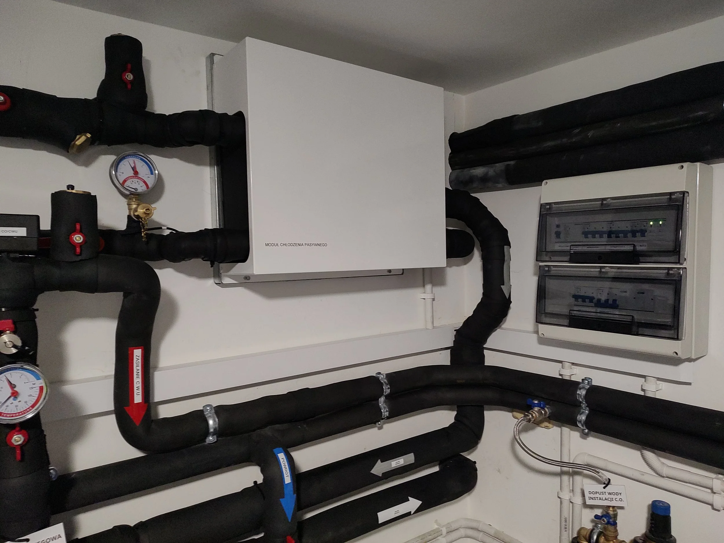 A mechanical room with black insulated pipes, gauges, valves, and an electrical panel.