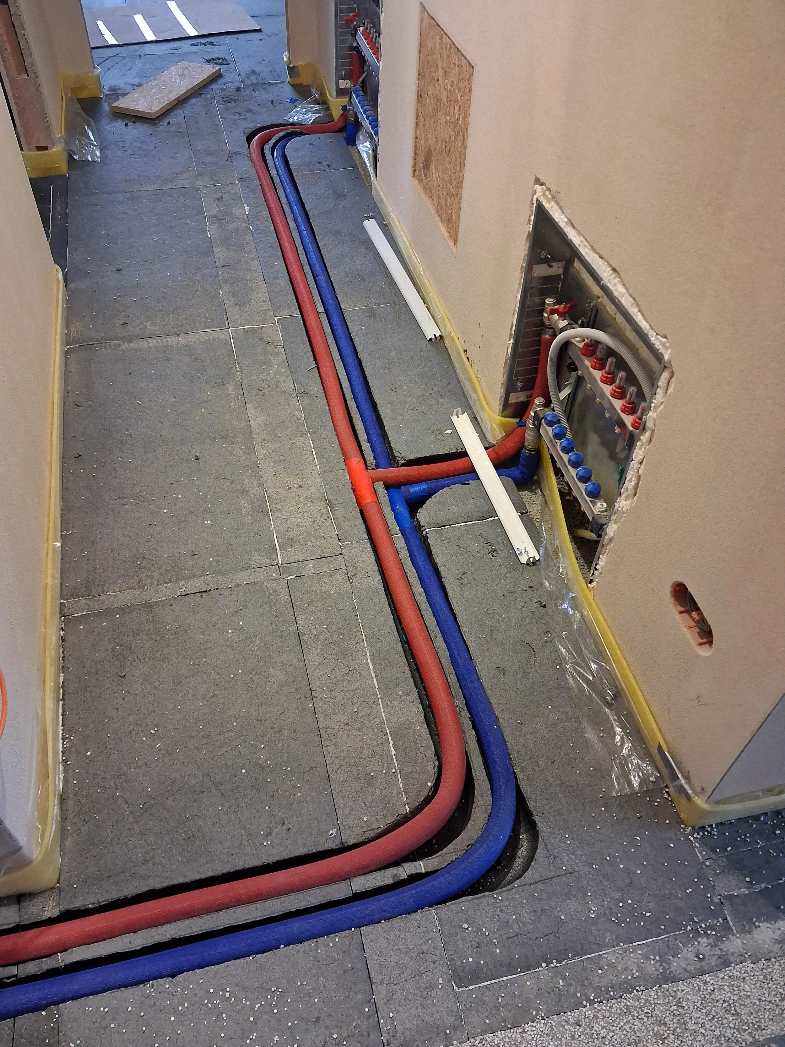 Construction area with exposed electrical wiring and plumbing pipes on a concrete floor, partially covered with protective plastic sheet, and a hole in the wall revealing electrical box and cabling.