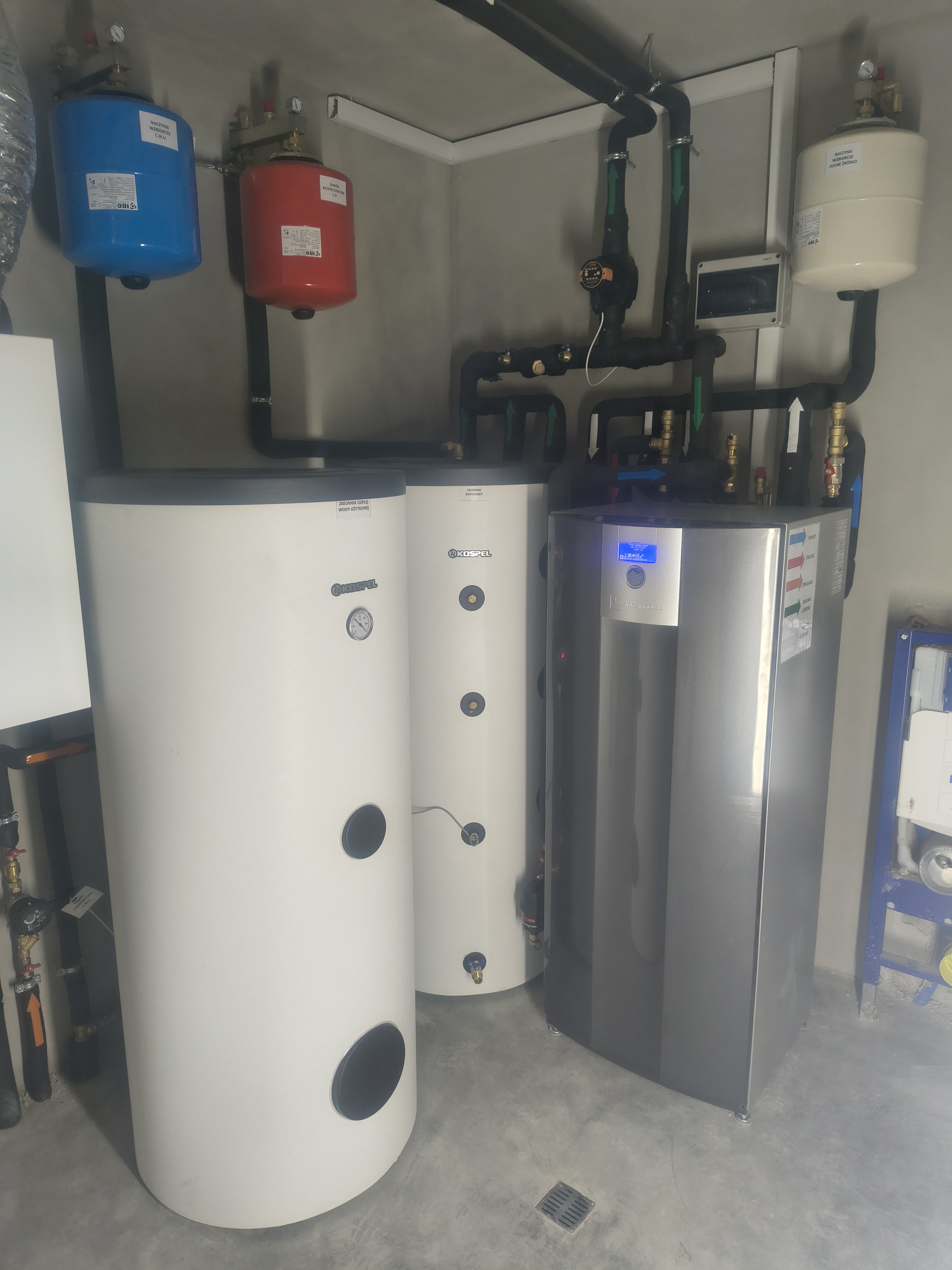 A utility room features a white water heater with a round shape, a control panel with digital display, and black and blue pipes with arrows indicating the flow direction. Nearby are red, blue, and white expansion tanks mounted on the wall.